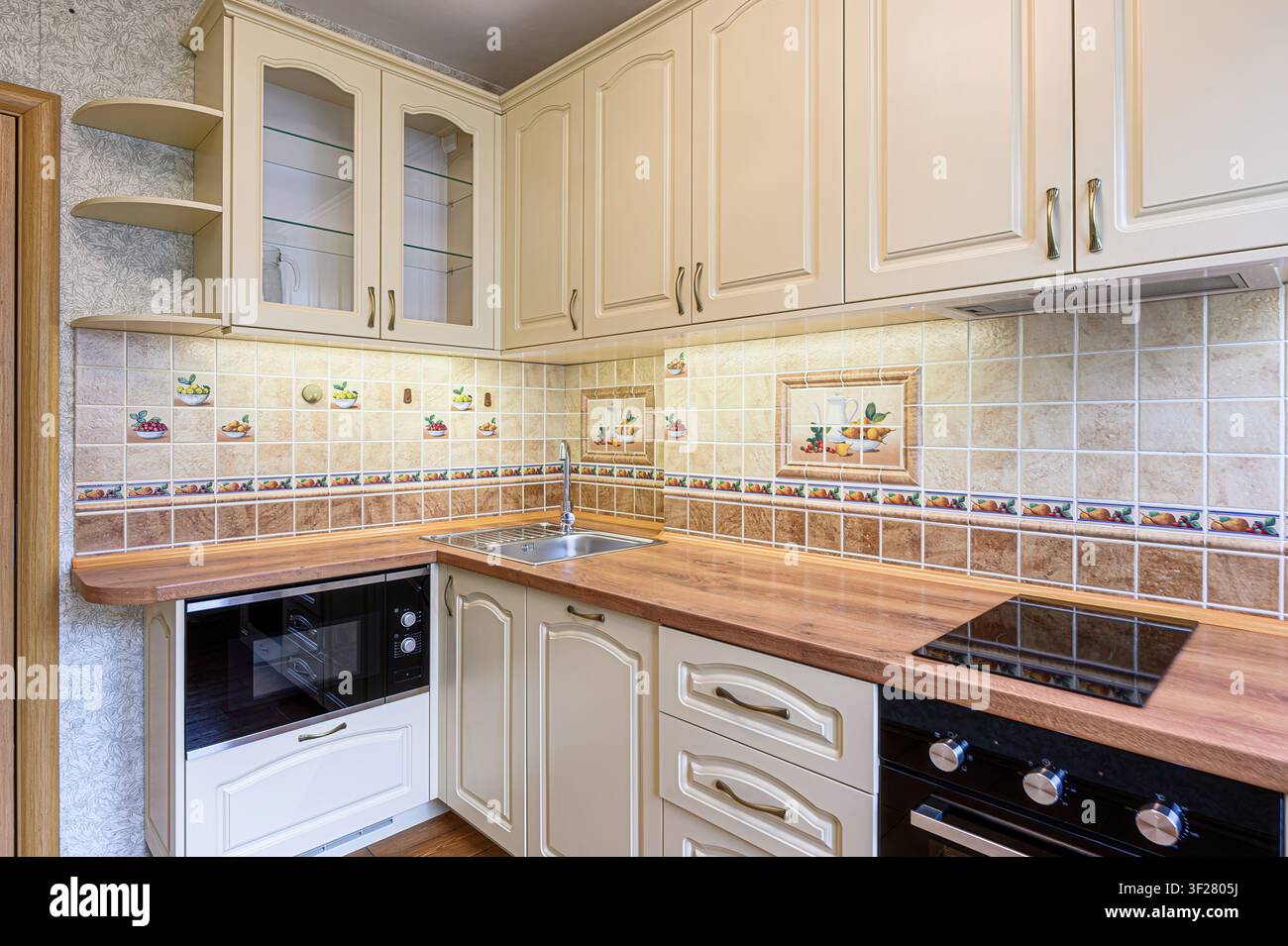 cucina ben illuminata con armadi color crema, un piano di lavoro in legno e un rivestimento decorativo in piastrelle Foto Stock