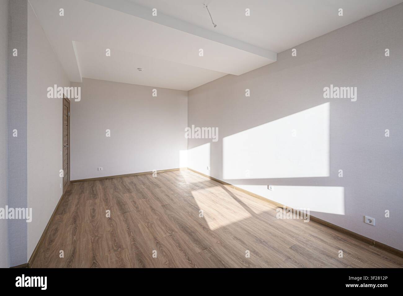 Una stanza vuota con pavimenti in legno e pareti bianche. La luce del sole entra creando forme luminose sulla parete. Una porta è visibile sul lato sinistro Foto Stock