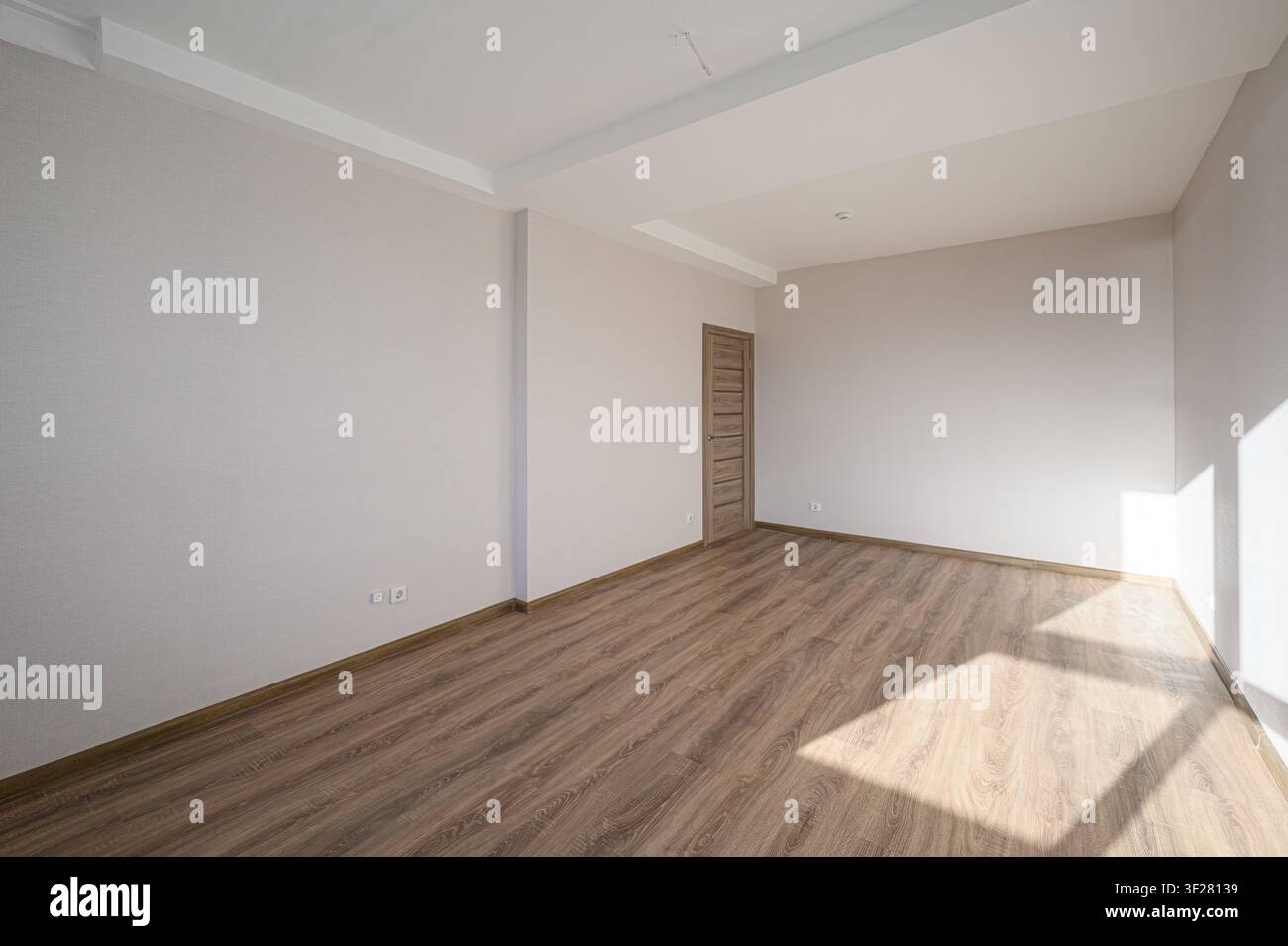 Una stanza vuota con pavimenti in legno e pareti bianche. La luce del sole entra creando forme luminose sulla parete. Una porta è visibile sul lato sinistro Foto Stock