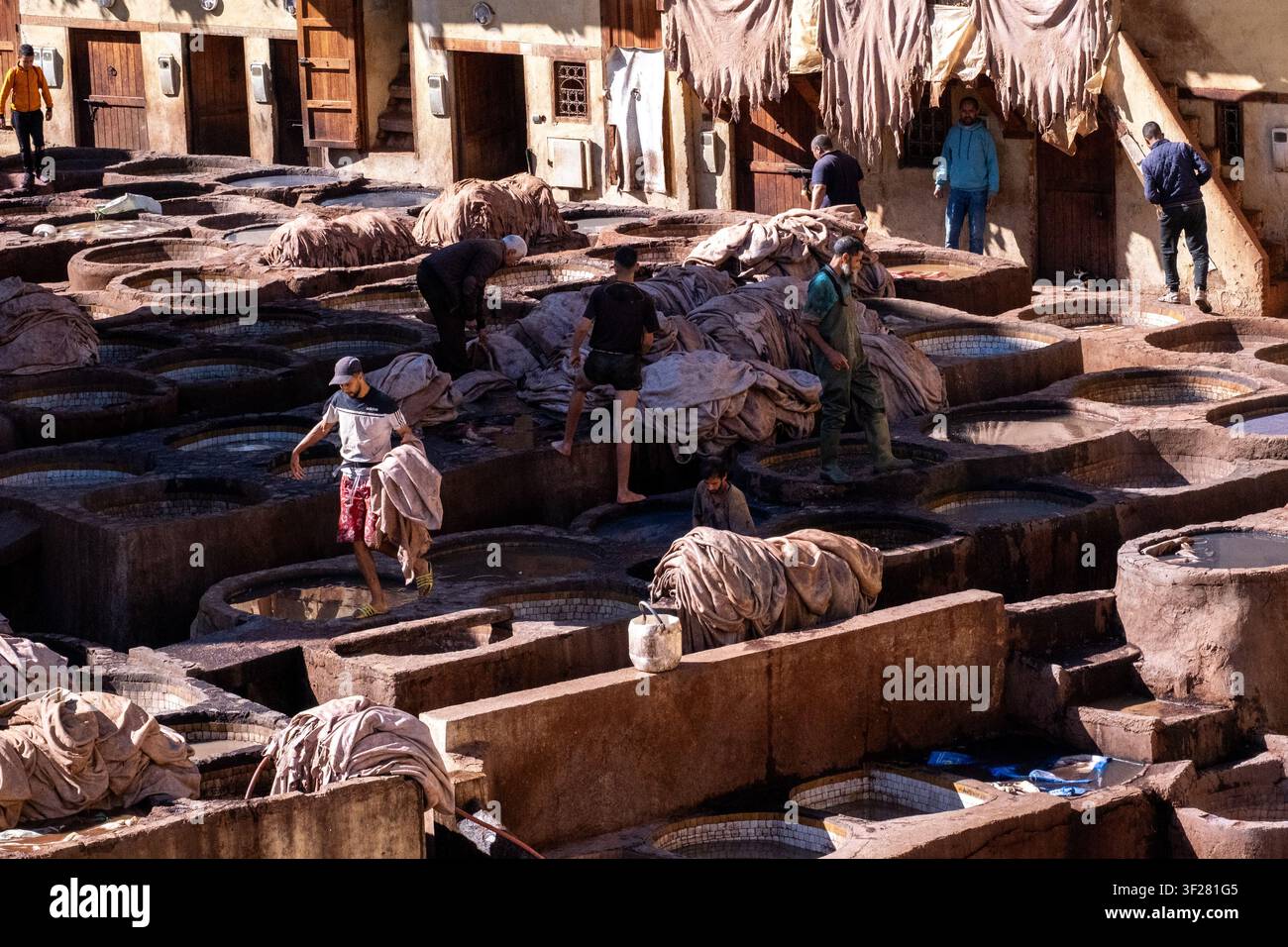 Vista ad angolazione alta di giorno di lavoratori che maneggiano pelli di cuoio bagnate tra vasche di concia rotonde, vecchie porte di legno, facciate in gesso e pelli in asciugatura a Chouara Foto Stock