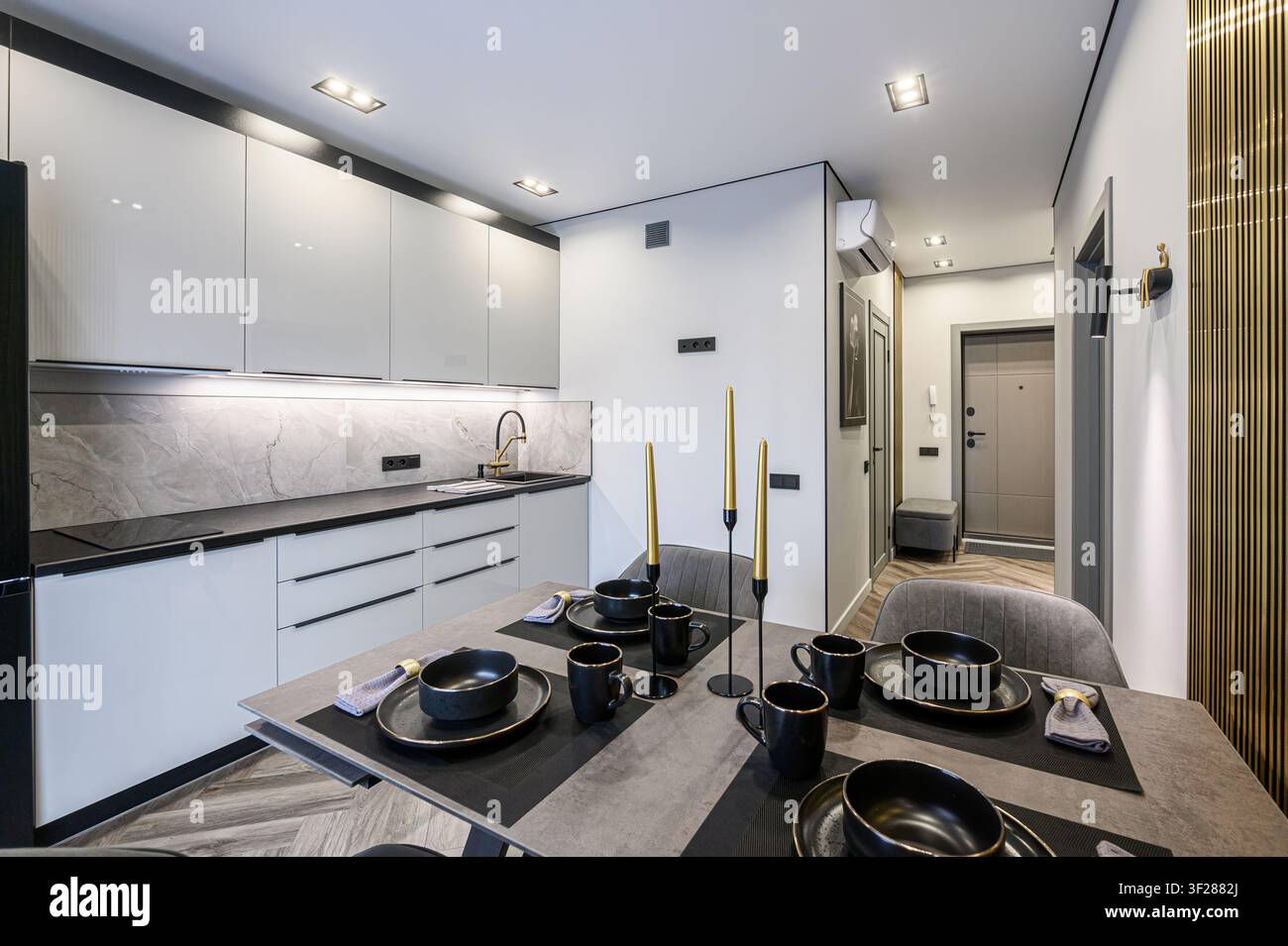 Cucina e sala da pranzo moderne. Armadi grigi, rivestimento in marmo, hardware nero e un tavolo apparecchiato con stoviglie nere. Sono visibili dettagli dorati Foto Stock