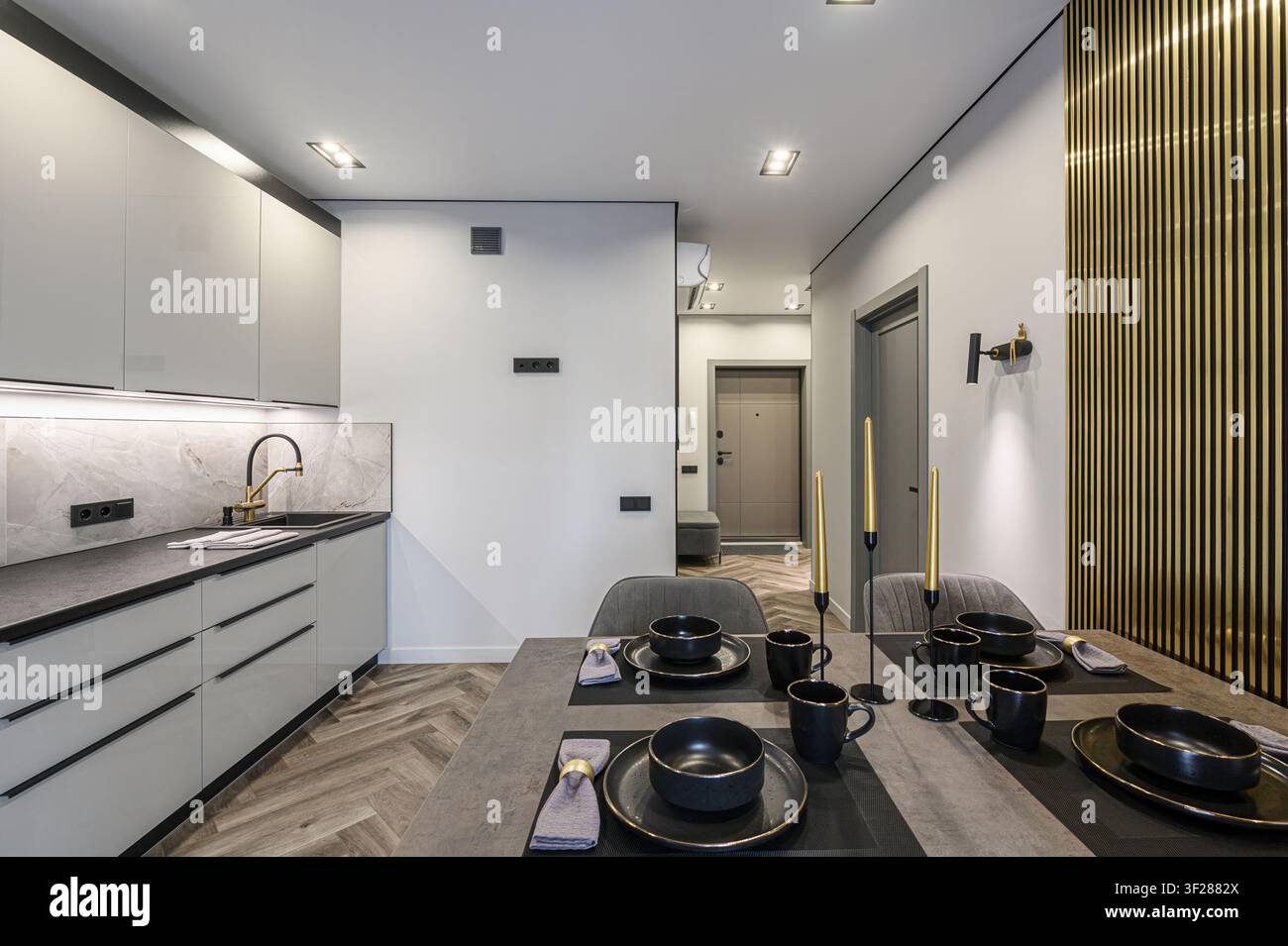Cucina e sala da pranzo moderne. Armadi grigi, rivestimento in marmo, hardware nero e un tavolo apparecchiato con stoviglie nere. Sono visibili dettagli dorati Foto Stock