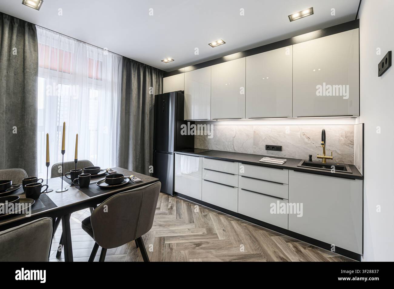 Cucina moderna con eleganti armadi bianchi, dettagli neri e una zona pranzo apparecchiata per un pasto. La luce naturale inonda lo spazio attraverso tende trasparenti Foto Stock