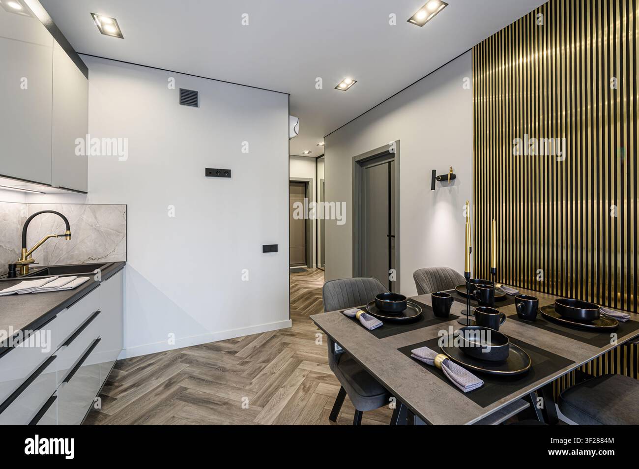 Cucina e sala da pranzo moderne. Armadi grigi, rivestimento in marmo, hardware nero e un tavolo apparecchiato con stoviglie nere. Sono visibili dettagli dorati Foto Stock