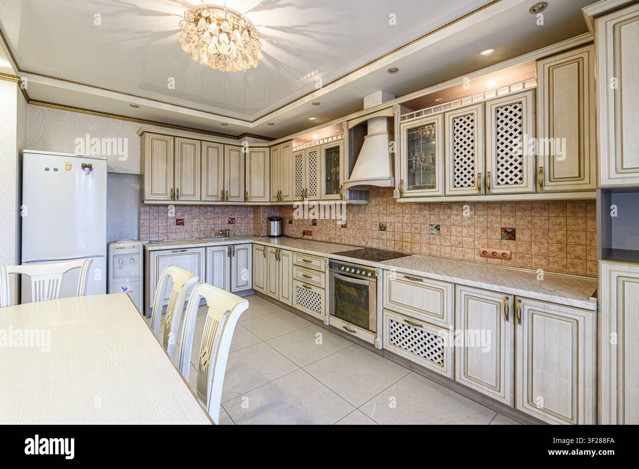 Una cucina ben illuminata presenta mobili color avorio con decorazioni dorate, un lampadario di cristallo e un pavimento piastrellato chiaro. Un frigorifero bianco è a sinistra, e una sala da pranzo Foto Stock