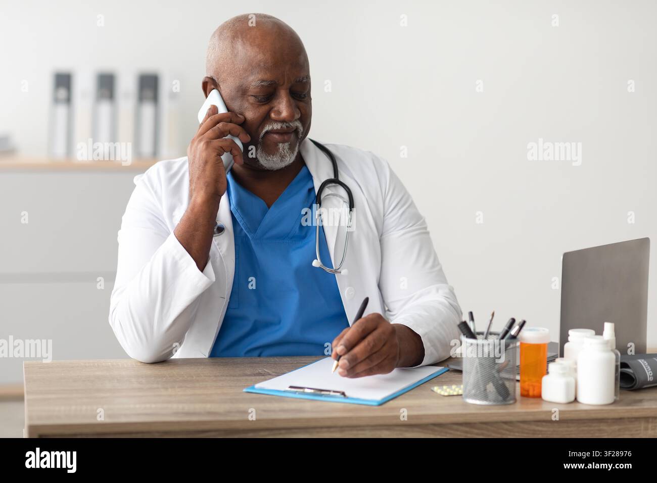 Medico uomo di colore che scrive appunti mentre parla al telefono in un ufficio sanitario Foto Stock