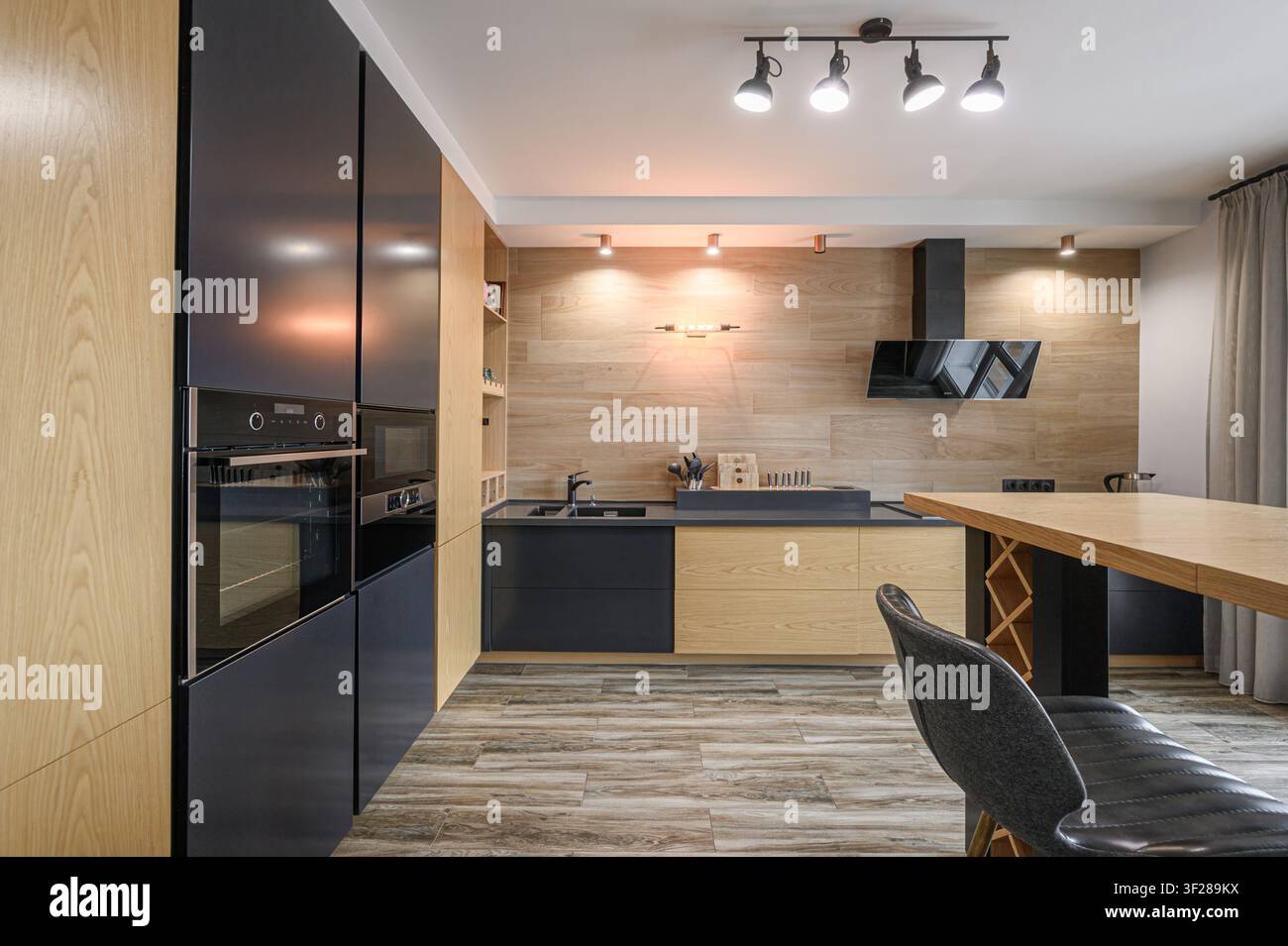 Cucina moderna con mobili in legno chiaro, dettagli neri e elettrodomestici in acciaio inossidabile. Ben illuminata con luci a binario, lo spazio presenta un retro in legno Foto Stock