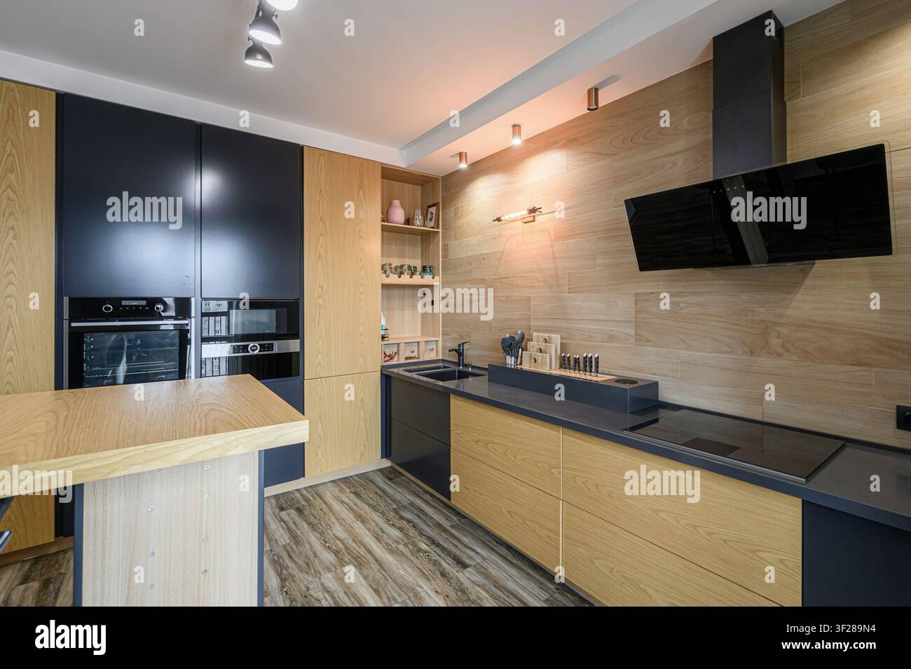 Cucina moderna con dettagli in legno, mobili neri e un piano di lavoro nero. Elettrodomestici eleganti e un'illuminazione stilosa completano il design Foto Stock