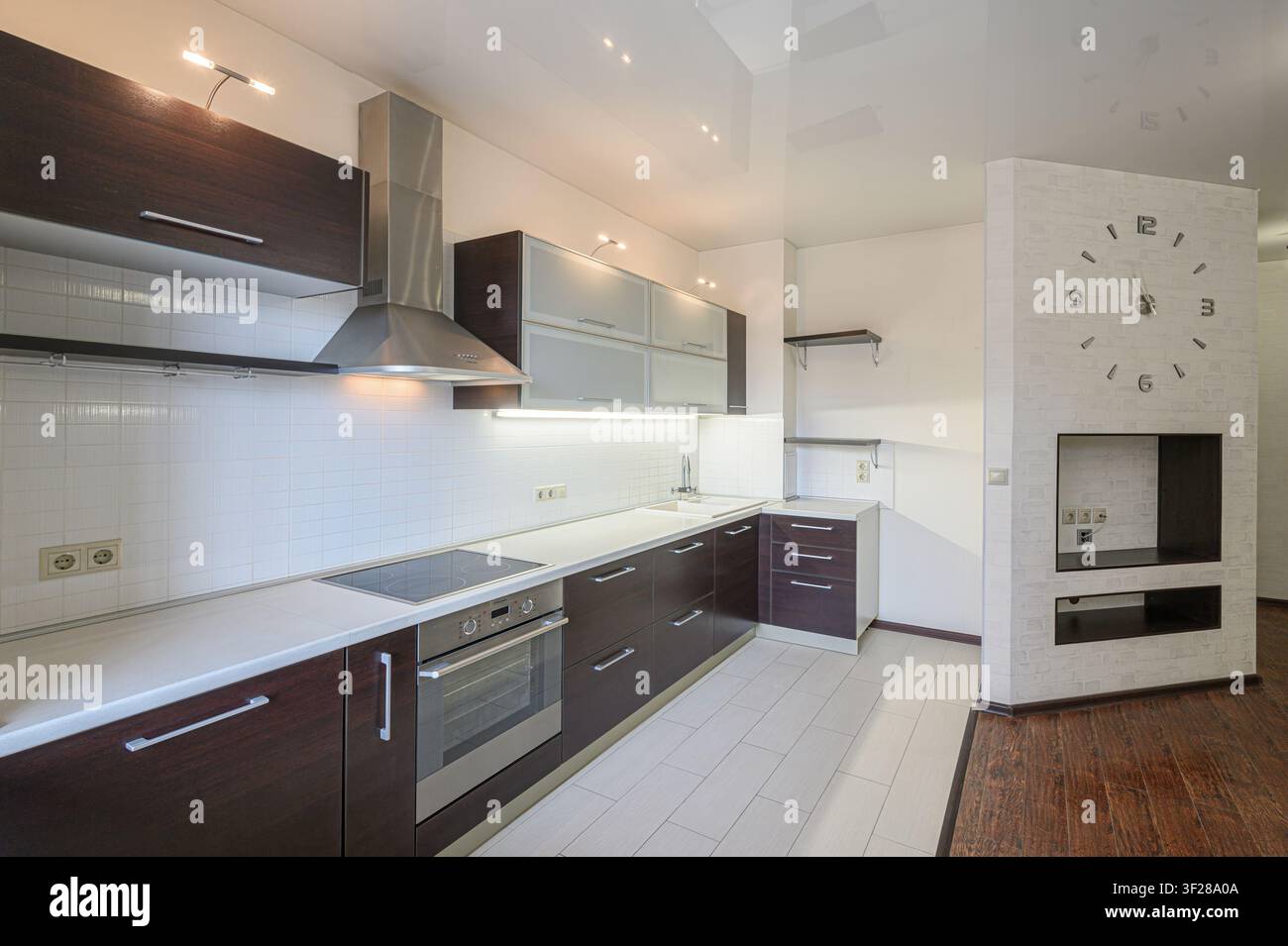 cucina moderna ben illuminata con armadi in legno scuro, piani di lavoro e piastrelle bianchi, elettrodomestici in acciaio inossidabile e pavimento in piastrelle chiare. Una grande Foto Stock