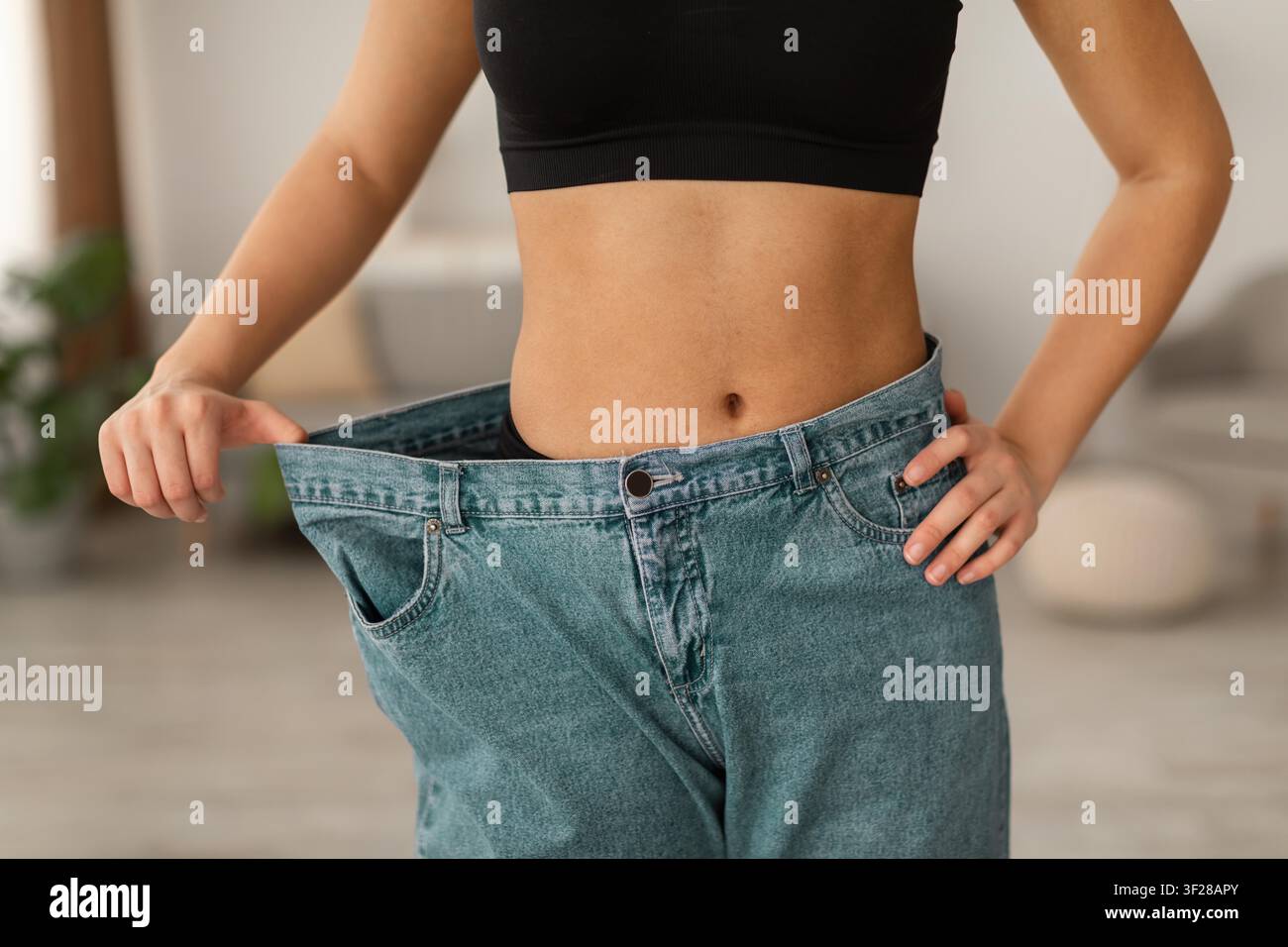 Donna con jeans oversize che celebra la perdita di peso, il percorso di fitness, ambientazione domestica Foto Stock