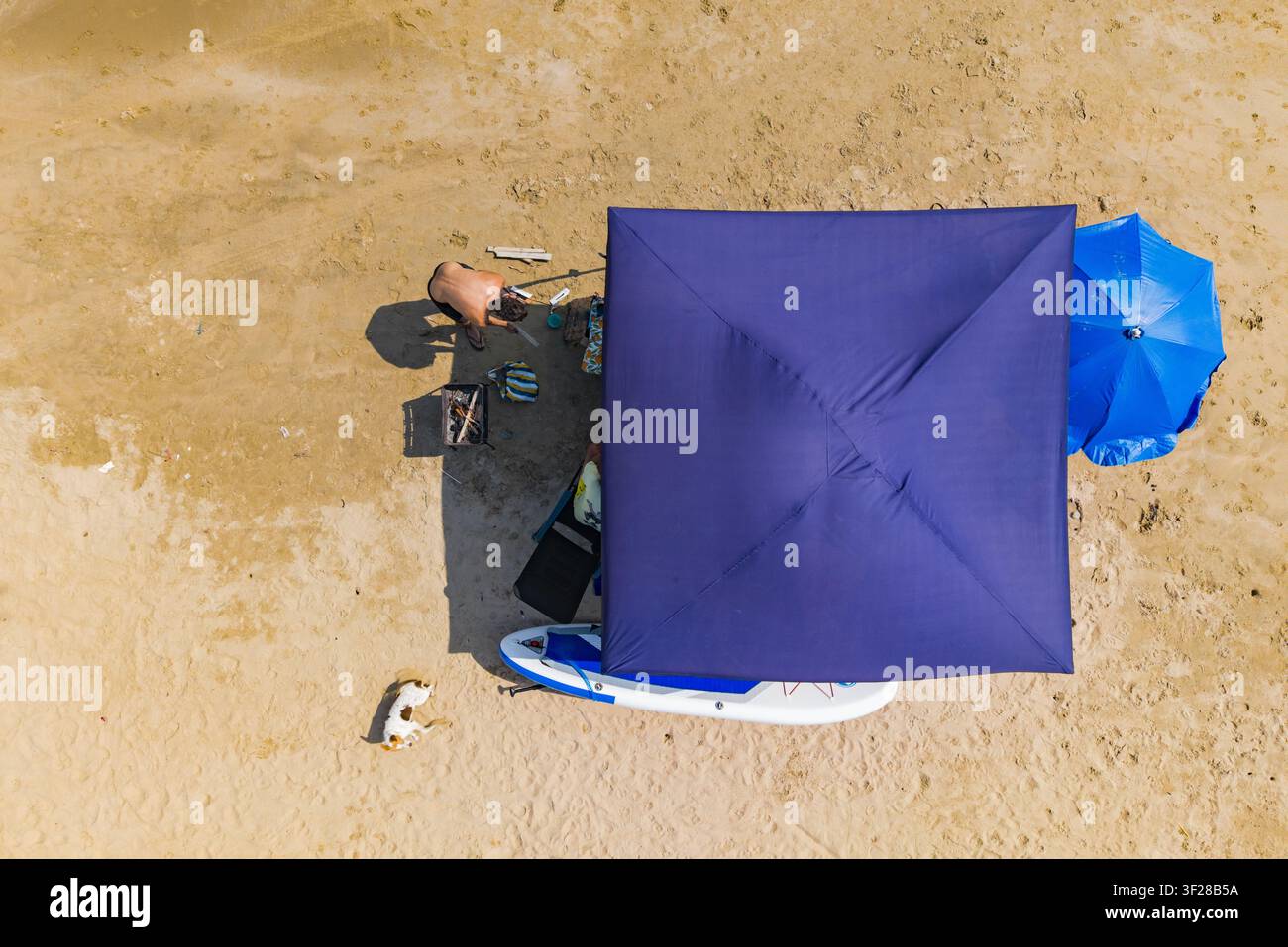 Vista aerea di una persona che prepara cibo su un grill accanto a un baldacchino blu, un ombrellone blu e una tavola da paddle sulla spiaggia sabbiosa di Ayangue, Santa Elena Foto Stock