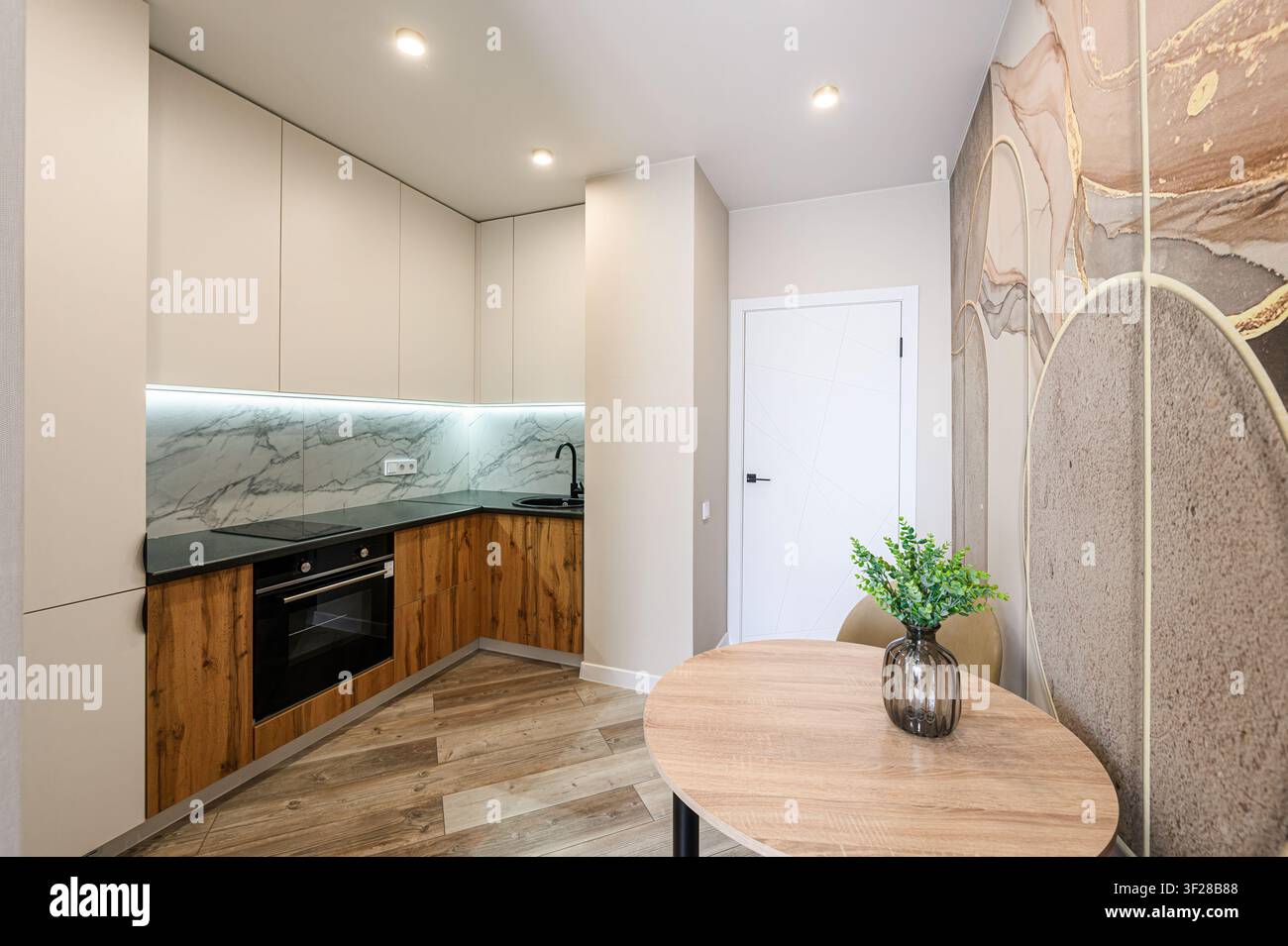 cucina moderna con armadi beige, dettagli in legno, un piano di lavoro nero, un retro in marmo e un tavolo rotondo in legno. L'arte astratta da parete aggiunge un tocco elegante Foto Stock