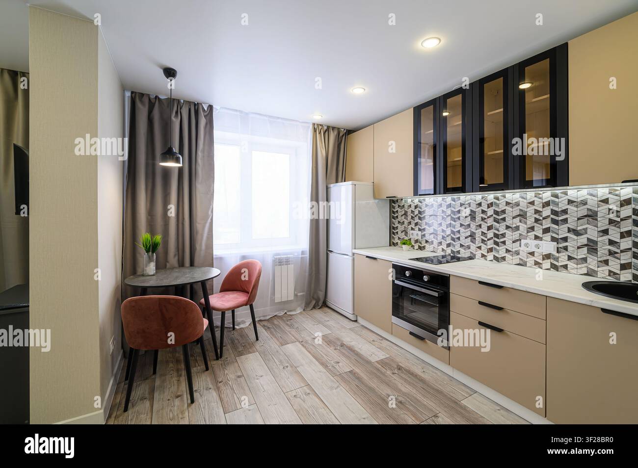 Cucina moderna con armadi beige, dettagli neri, un tavolo da pranzo con due sedie rosa, pavimenti effetto legno e piastrelle geometriche per il rivestimento murale Foto Stock