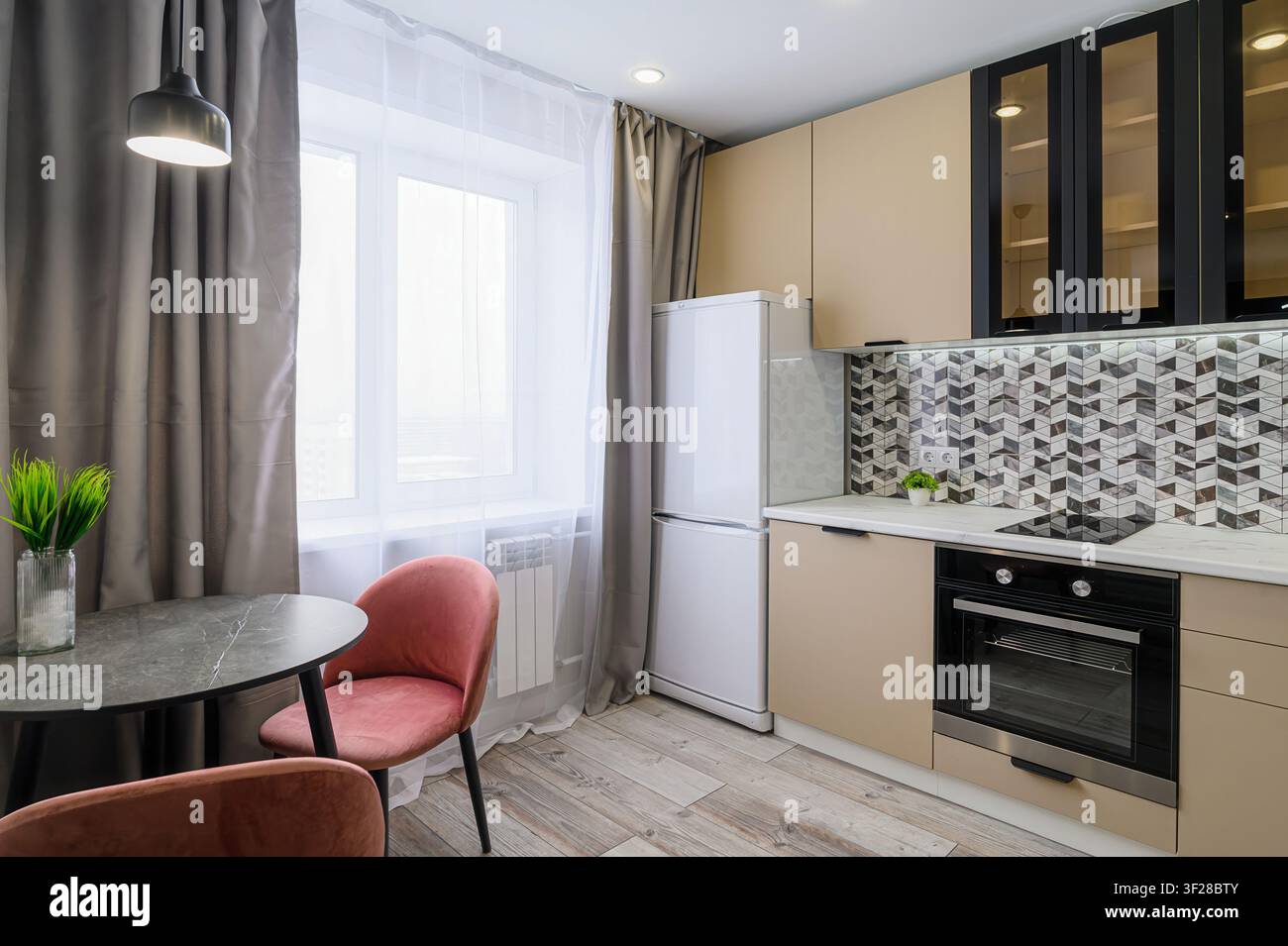 cucina accogliente con armadi beige, dettagli neri, un rivestimento a mosaico e un piccolo tavolo rotondo con sedie rosa. Un frigorifero bianco si trova vicino a una finestra wi Foto Stock