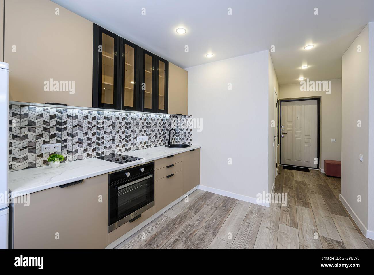 Cucina moderna con armadi beige, dettagli neri, schiera geometrica e pavimento effetto legno che conduce a un corridoio con una porta Foto Stock
