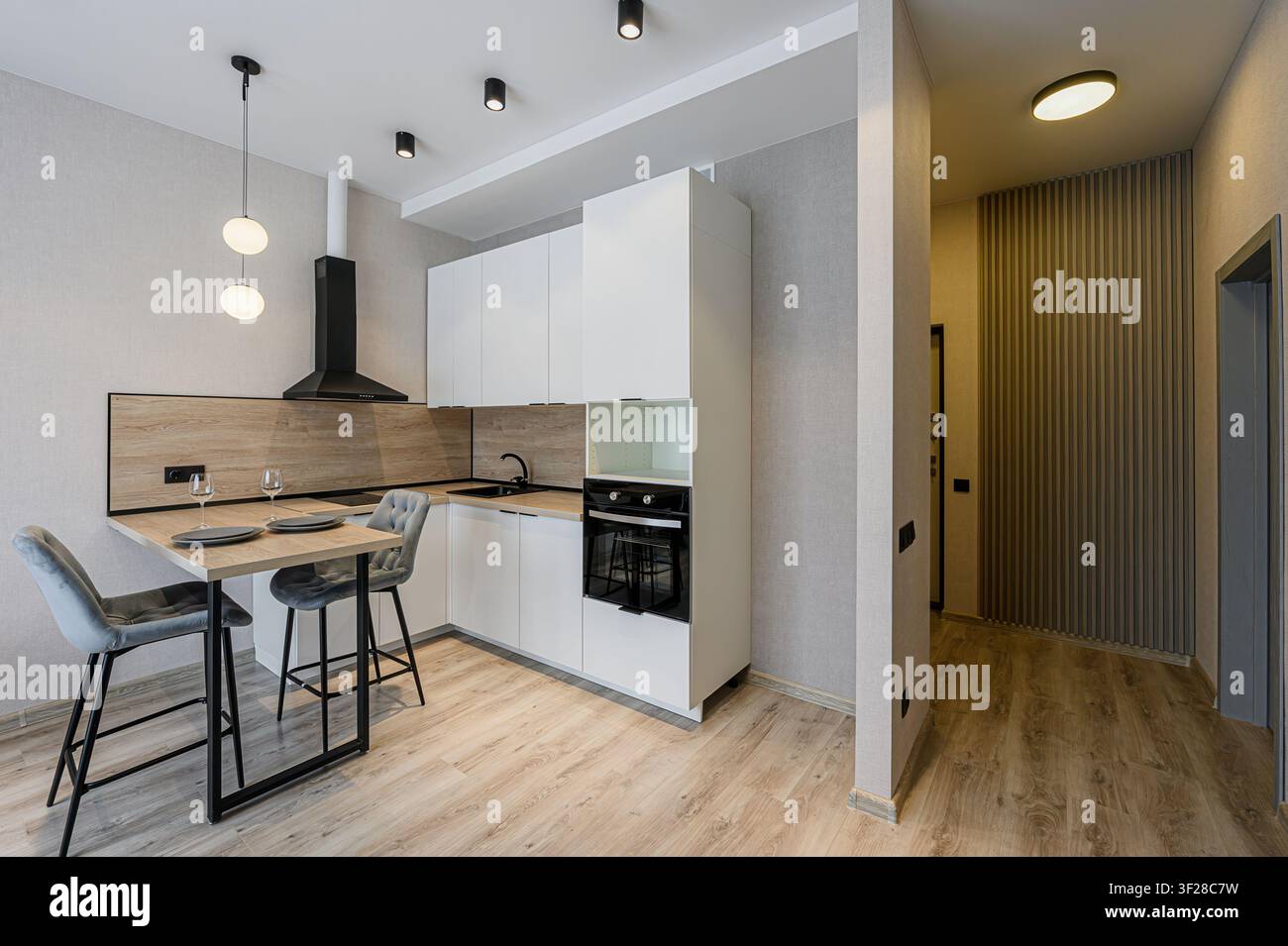 cucina moderna con armadi bianchi, una cappa nera, dettagli in legno e un tavolo da pranzo con due sedie. Un corridoio è visibile a destra Foto Stock