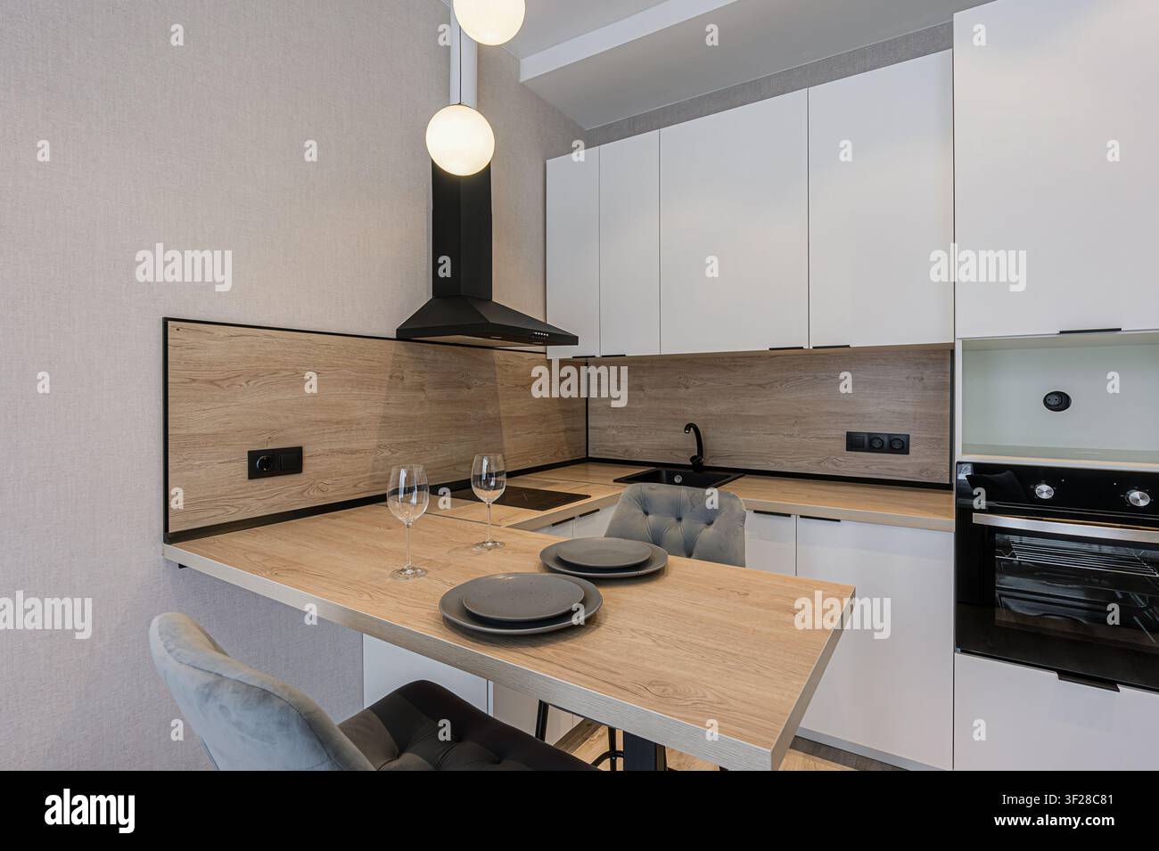 Cucina moderna con armadi bianchi, dettagli in legno, elettrodomestici neri e un bancone della colazione con sedie grigie. Bicchieri e piatti sono disposti sul bancone Foto Stock