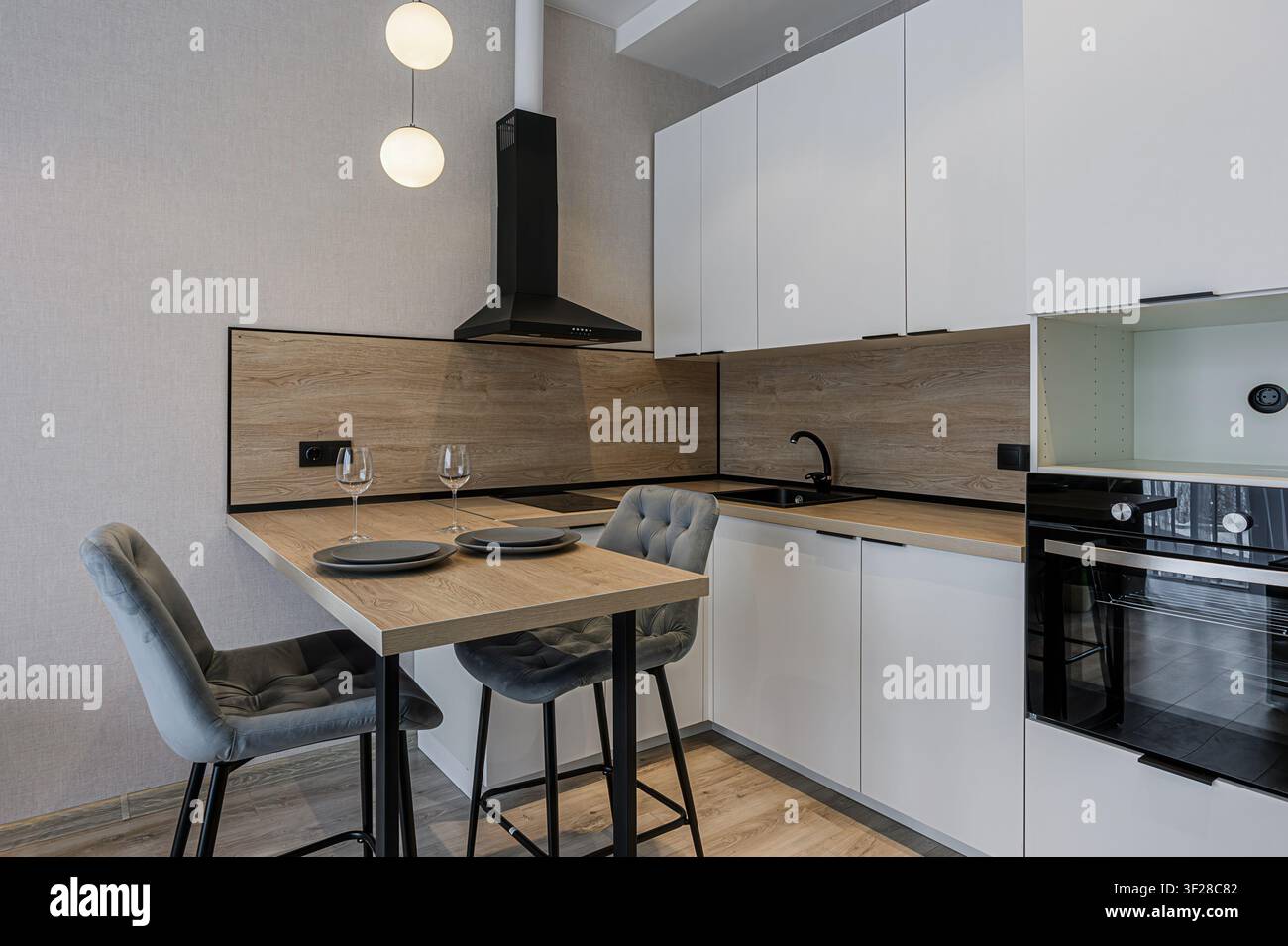 cucina moderna con armadi bianchi, dettagli in legno, rubinetteria nera e sedie grigie a un piccolo tavolo da pranzo. Lampade a sospensione a globo aggiungono un tocco elegante Foto Stock