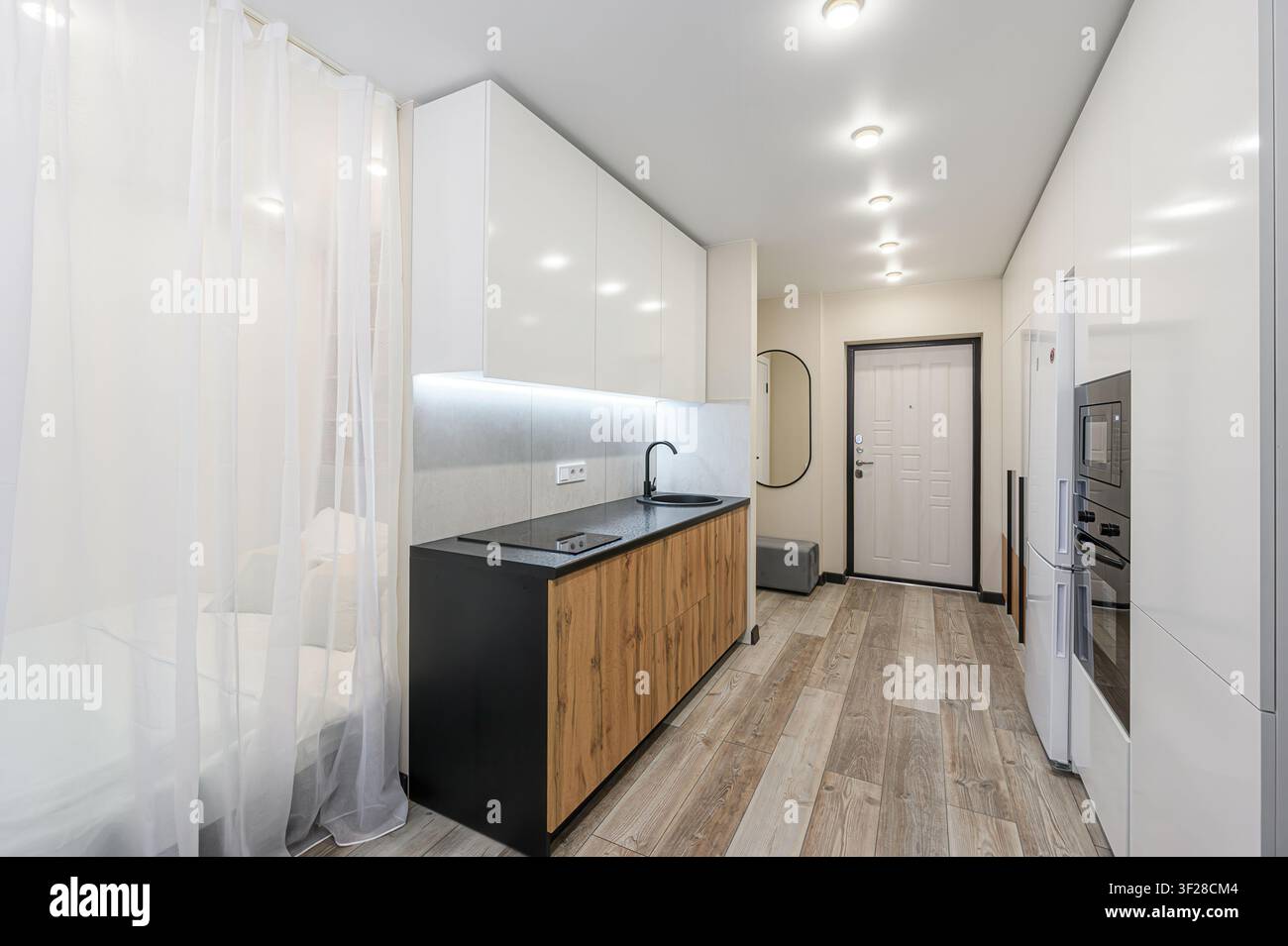 appartamento moderno ben illuminato con una cucina compatta con eleganti armadi bianchi, un piano di lavoro nero e dettagli in legno. Tende trasparenti separano il Foto Stock