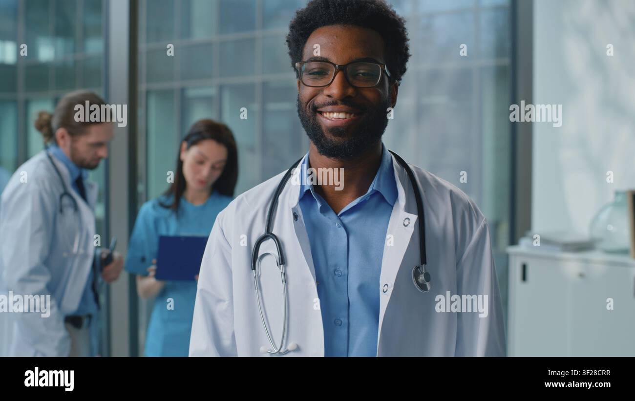 Uomo afroamericano medico che pensa, guarda la telecamera, sorride con un sorriso ampio e felice in un laboratorio clinico vicino a lavoratori della medicina sullo sfondo. Uomo etnico Foto Stock