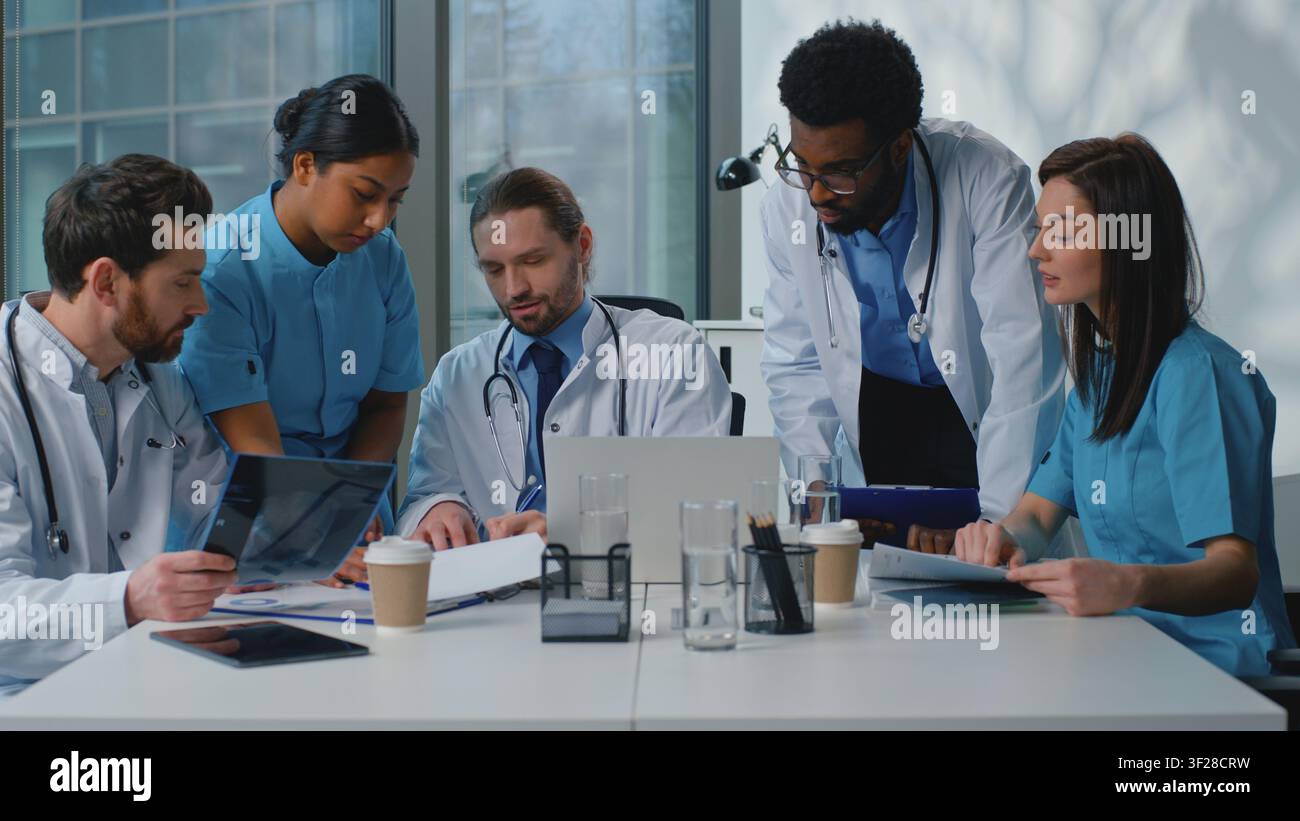 Gruppo serio di medici multirazziali che discutono a tavola e lavorano insieme in clinica. Leader caucasico dà consigli agli internisti, team di assistenza, colleghi Foto Stock