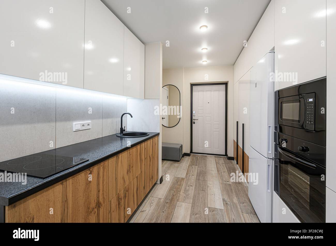 Cucina moderna e corridoio con mobili bianchi, dettagli in legno, elettrodomestici neri e pavimentazione effetto legno. Un'illuminazione luminosa illumina lo spazio Foto Stock