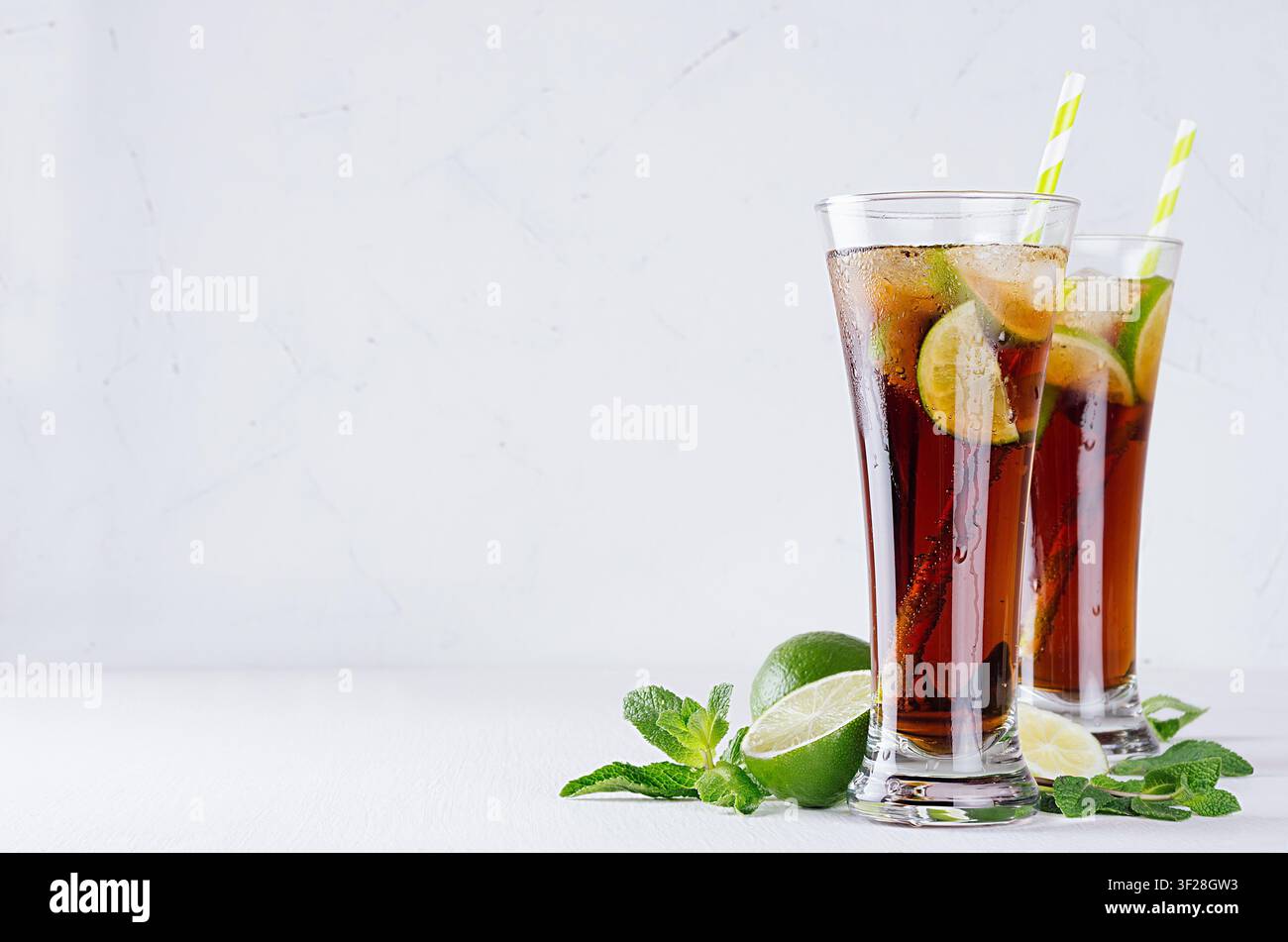 Cocktail freddo cuba libre in due lunghi bicchieri bagnati con paglia, pezzi di lime, menta su morbido sfondo bianco moderno. Copia spazio. Foto Stock