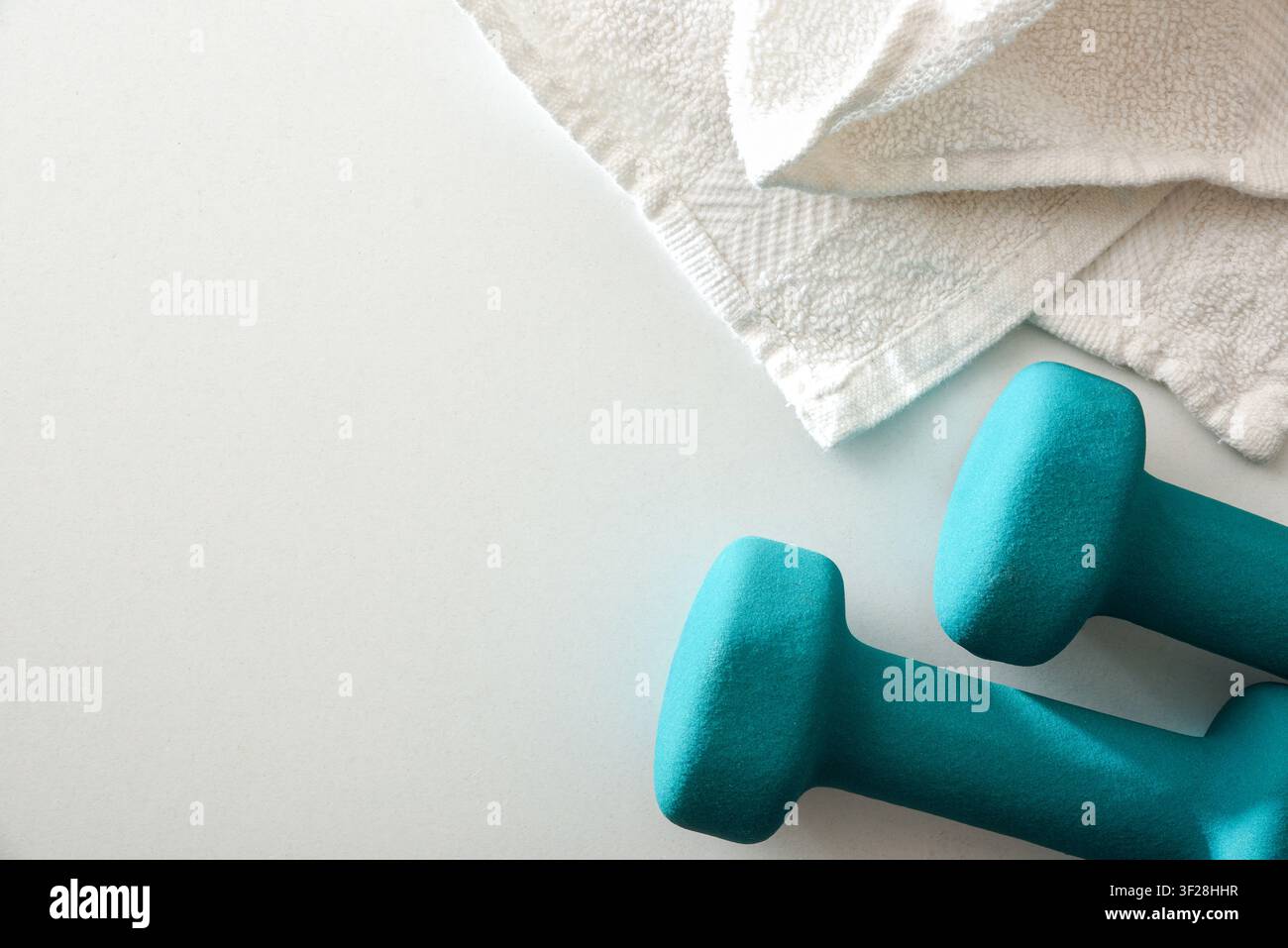 Due manubri in neoprene turchesi accanto a un asciugamano bianco su una superficie luminosa con luce naturale, rappresentanti fitness, allenamenti a casa, forza e salute Foto Stock