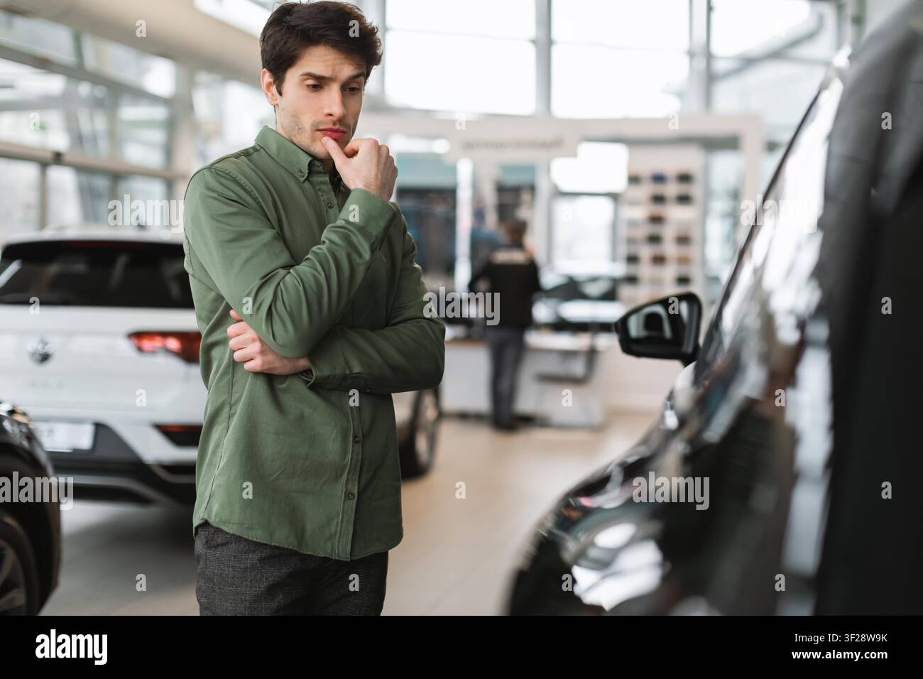 Giovane uomo caucasico pensieroso che considera l'acquisto di un'auto in una sala espositiva, concetto aziendale Foto Stock