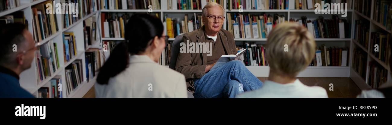 Testa di uomo caucasico anziano che conduce una discussione di gruppo leggendo un libro in biblioteca, sostenendo l'apprendimento degli adulti e l'istruzione comunitaria. Piano ampio, ideale per campagne di alfabetizzazione o workshop Foto Stock