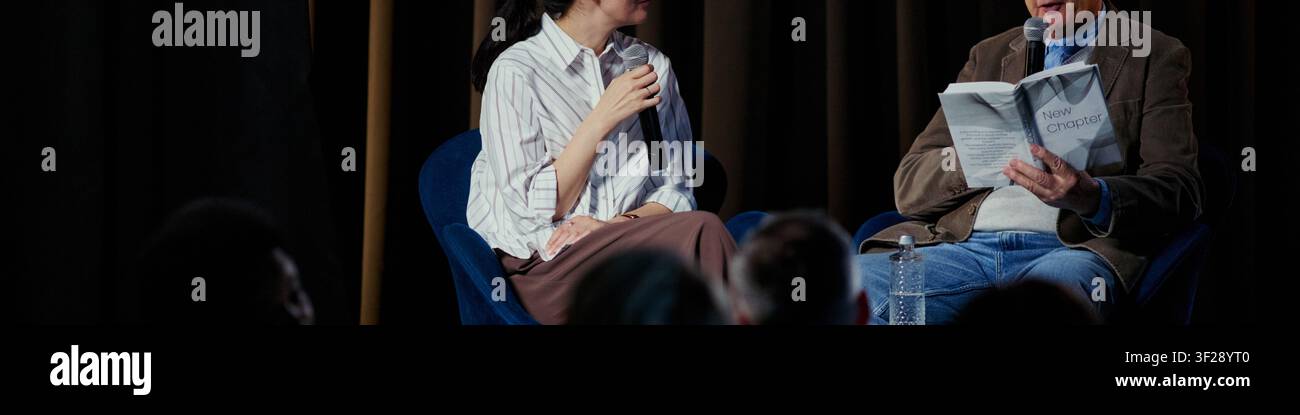Intestazione di una giovane donna e un uomo anziano che parlano nei microfoni durante una discussione pubblica, condividendo idee e leggendo appunti. Utile per il marketing di conferenze, eventi educativi, promozione di panel Foto Stock