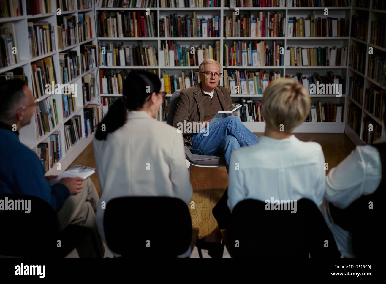 Uomo anziano caucasico che conduce una discussione di gruppo in biblioteca, tiene un libro e guida i partecipanti adulti in una sessione di apprendimento. Utile per educazione, coaching, club del libro, laboratorio comunitario Foto Stock