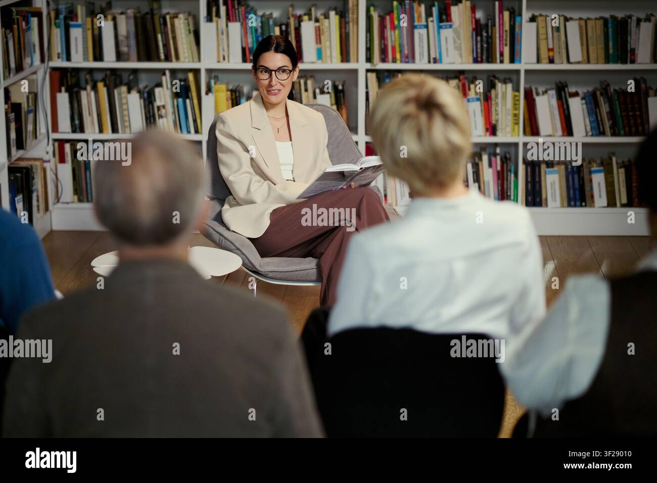 Donna caucasica di mezza età che conduce una discussione su un libro e legge a un gruppo di adulti in biblioteca, sostenendo l'istruzione e l'apprendimento comunitario. Scatto ampio, ideale per programmi di alfabetizzazione o workshop Foto Stock