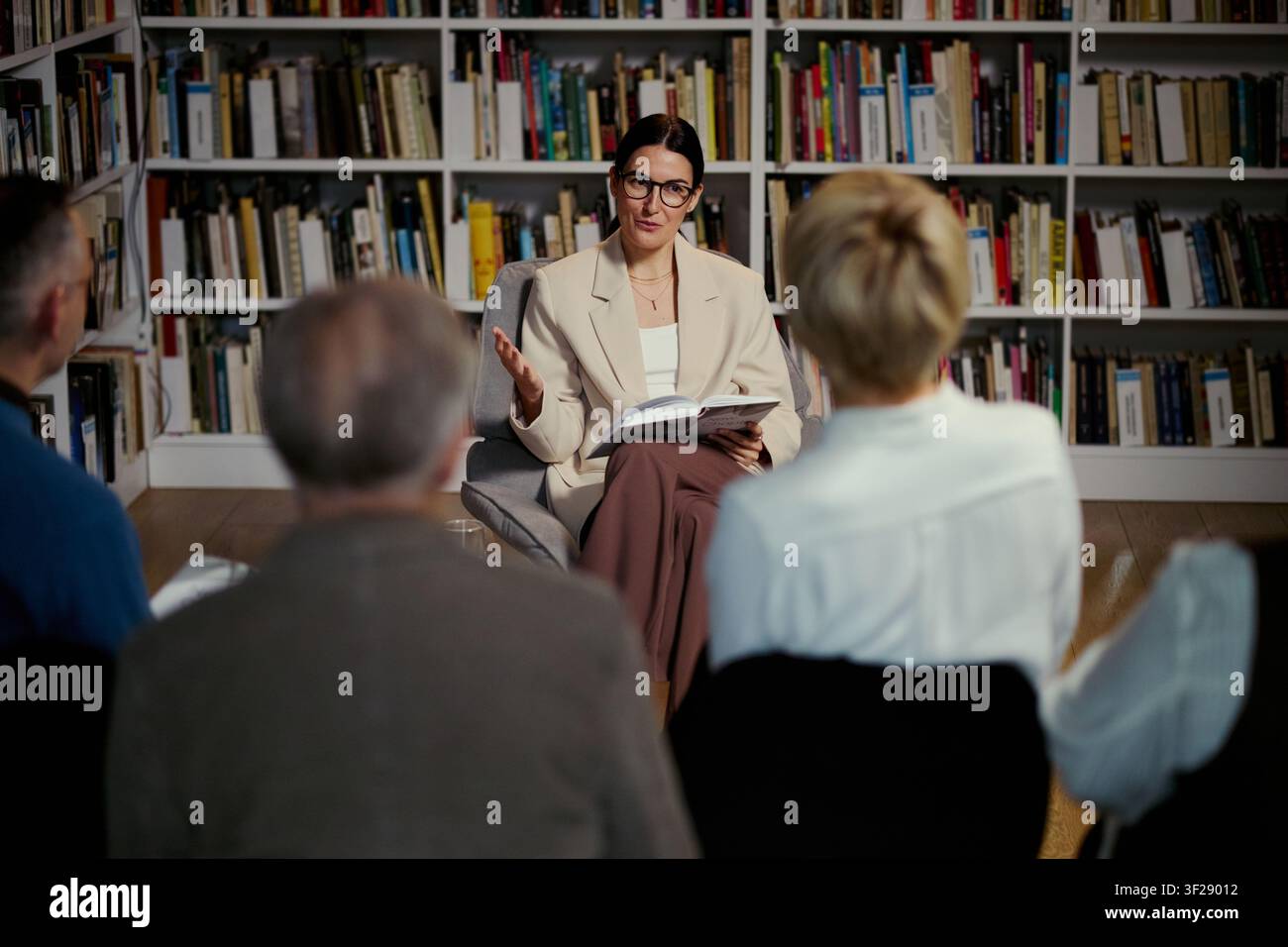 Donna caucasica di mezza età che conduce una discussione di gruppo tenendo un libro in biblioteca, sostenendo l'apprendimento degli adulti e la comunicazione. Utile per l'istruzione, il coaching, il marketing di workshop Foto Stock