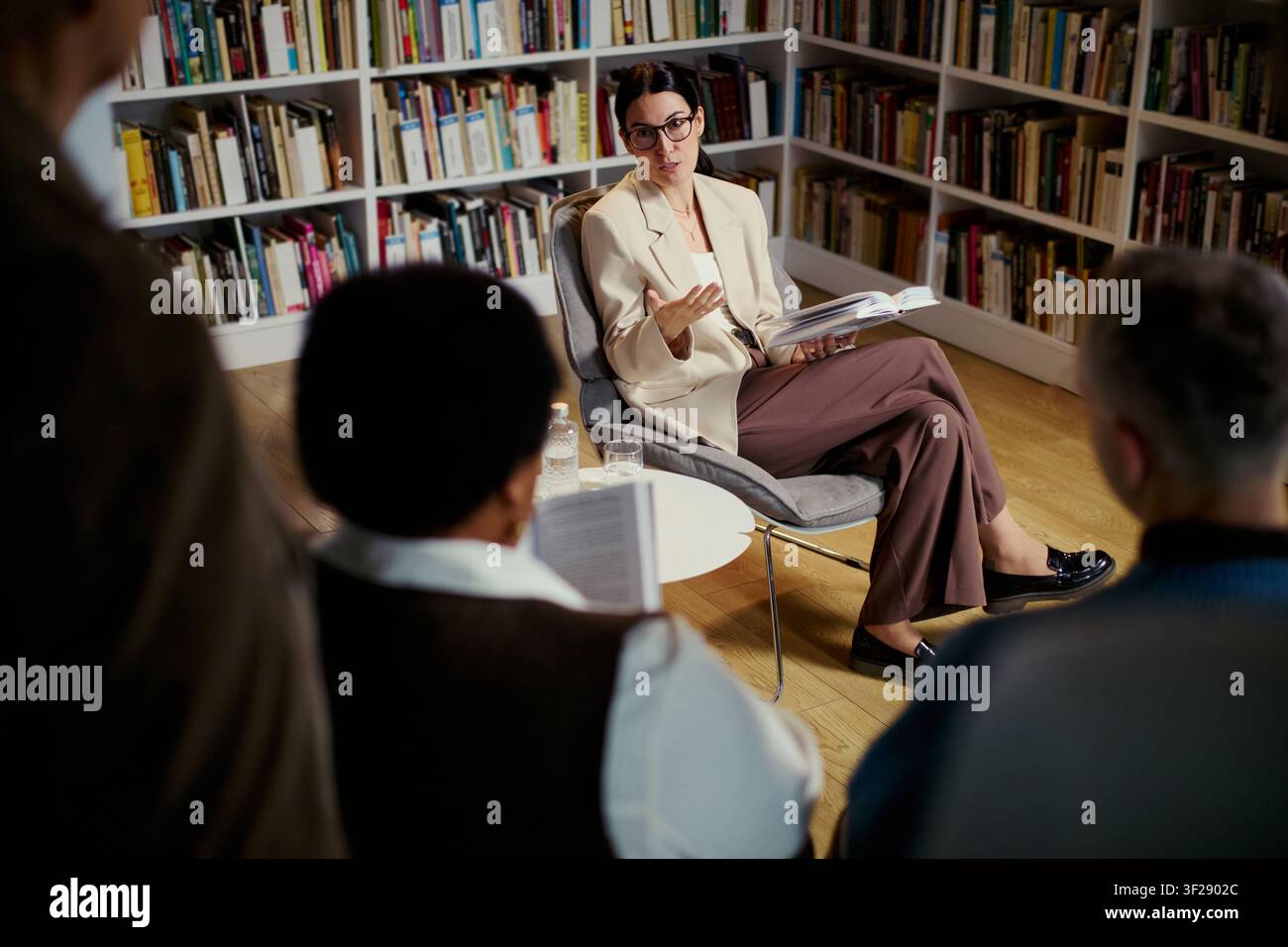 Giovane donna caucasica che conduce una discussione su un libro, gesticolando mentre legge a un piccolo gruppo in biblioteca, sostenendo i temi dell'educazione e della comunicazione. Inquadratura ampia, ideale per campagne di apprendimento Foto Stock