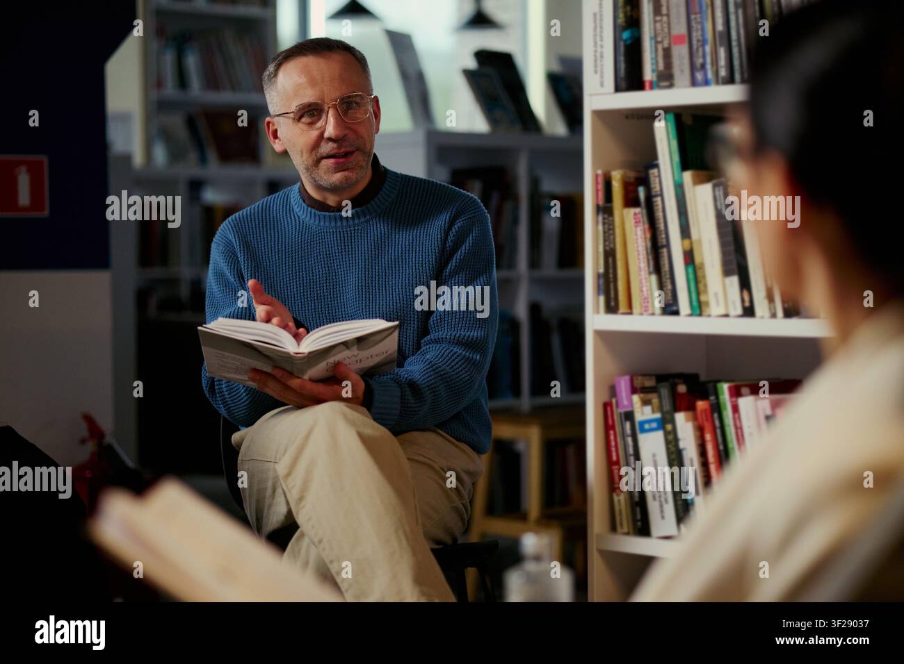 Uomo di mezza età caucasico che legge un libro e guida una discussione in biblioteca, sostenendo l'apprendimento degli adulti e l'educazione di gruppo. Utile per programmi di alfabetizzazione, workshop, club del libro Foto Stock