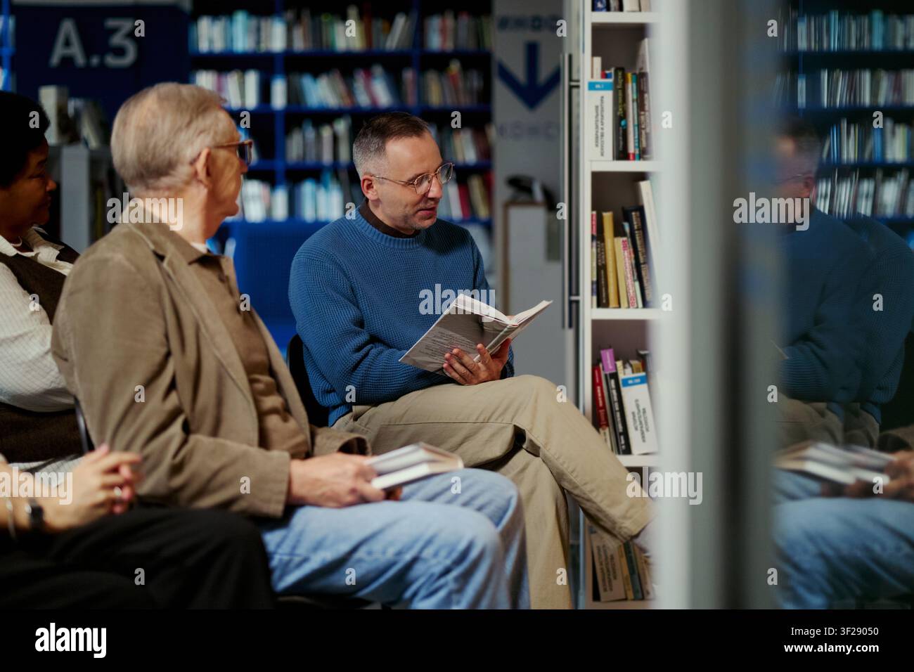 Uomo di mezza età che legge un libro e guida una discussione con adulti diversificati in biblioteca, sostenendo l'apprendimento comunitario e l'alfabetizzazione. Utile per programmi educativi e promozione di club del libro Foto Stock