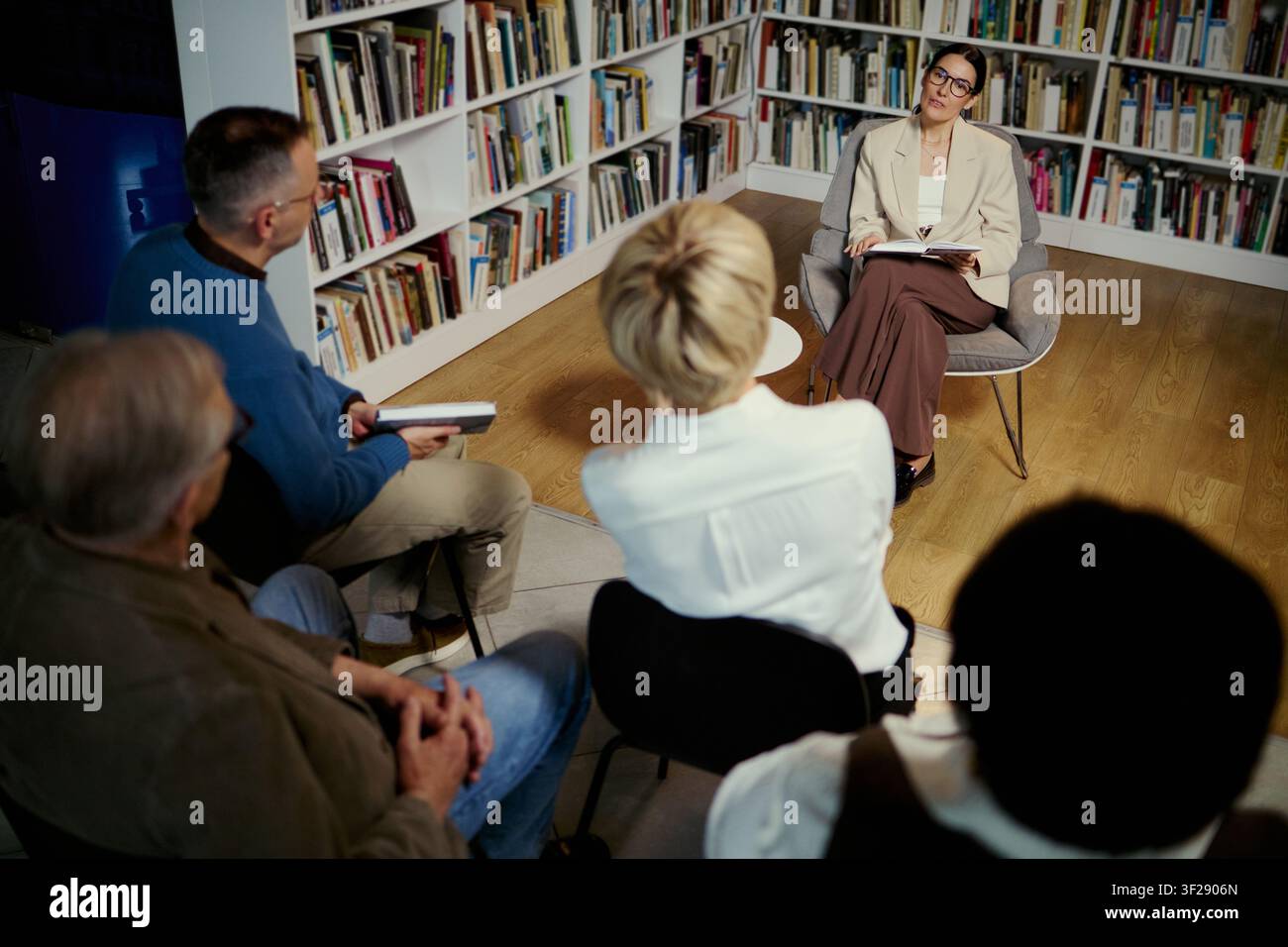 Vista ad angolo alto di una donna caucasica di mezza età che conduce una seduta di terapia di gruppo, ascoltando adulti in discussione in biblioteca. Ideale per la salute mentale, consulenza, marketing di programmi di supporto Foto Stock