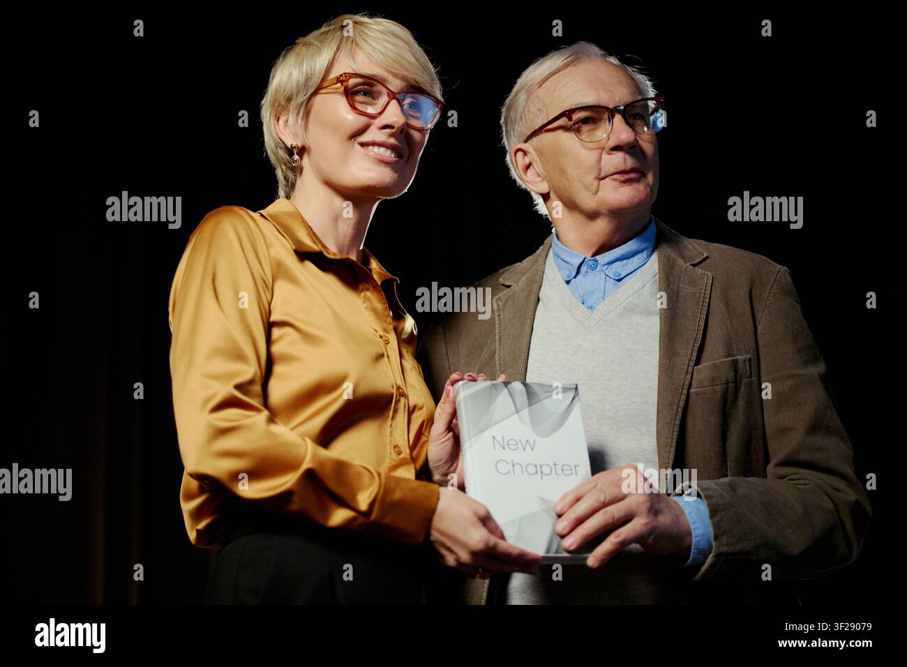 Uomo anziano caucasico e donna di mezza età che tengono un libro sul palco, segnando una transizione di carriera e una pietra miliare del mentorato. Utile per pubblicazioni, pianificazione della pensione, campagne di leadership Foto Stock