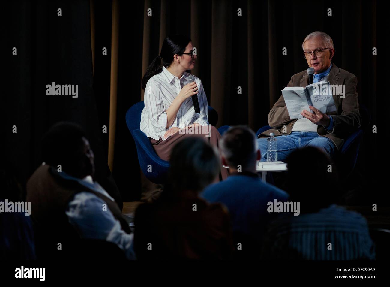 Inquadratura ampia di un uomo caucasico anziano che legge da un libro mentre una giovane donna modera una discussione sul palco davanti al pubblico. Ideale per la pubblicazione di eventi, incontri con autori, marketing della public speaking Foto Stock