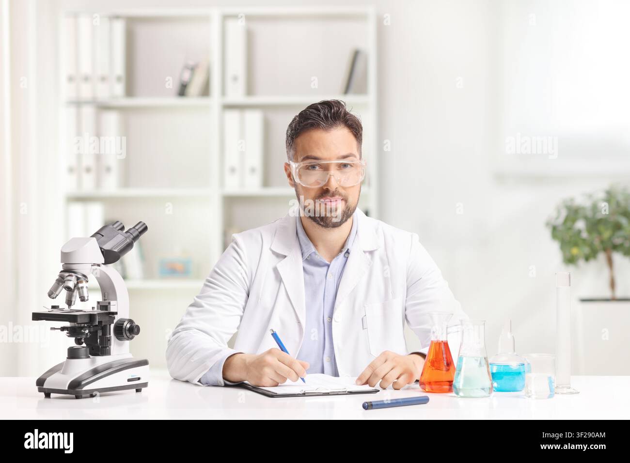 Scienziato seduto a una scrivania con un microscopio e fiale di sostanze chimiche in un ufficio Foto Stock