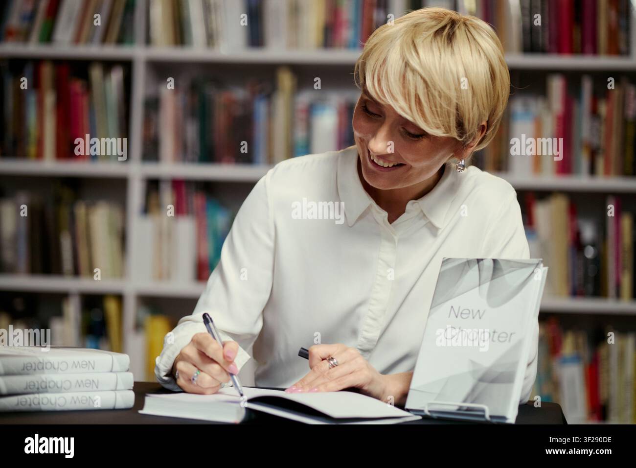 Giovane donna caucasica che autografa un libro a un tavolo di libreria, sorridente durante la promozione di una nuova uscita. Utile per il marketing editoriale, il branding degli autori, gli eventi letterari, primo piano Foto Stock