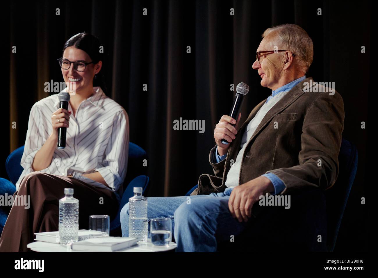 Giovane donna adulta e uomo anziano che parlano nei microfoni durante una discussione sul palco, presentando intuizioni sulla leadership e sulla comunicazione pubblica. Ideale per eventi aziendali, marketing di panel Foto Stock