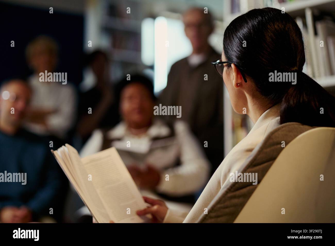 Giovane donna asiatica che legge un libro a un gruppo diversificato durante una discussione, mostrando educazione e apprendimento comunitario. Utile per programmi di alfabetizzazione, club del libro, workshop, primo piano Foto Stock