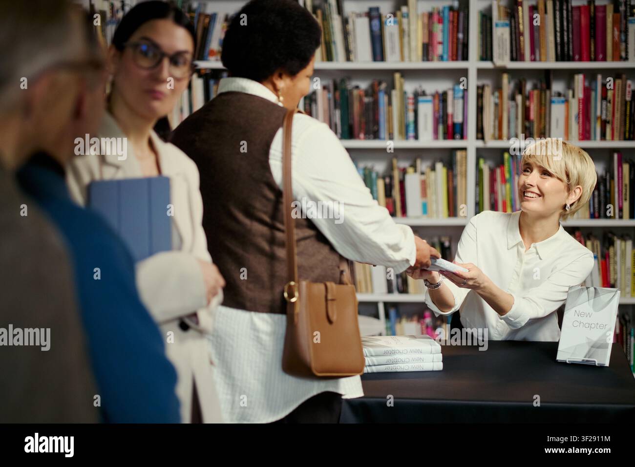 Donna caucasica di mezza età che autografa un libro e saluta un lettore durante un evento in libreria, con una fila in attesa nelle vicinanze. Ideale per il marketing editoriale, la promozione degli autori, il coinvolgimento della comunità Foto Stock