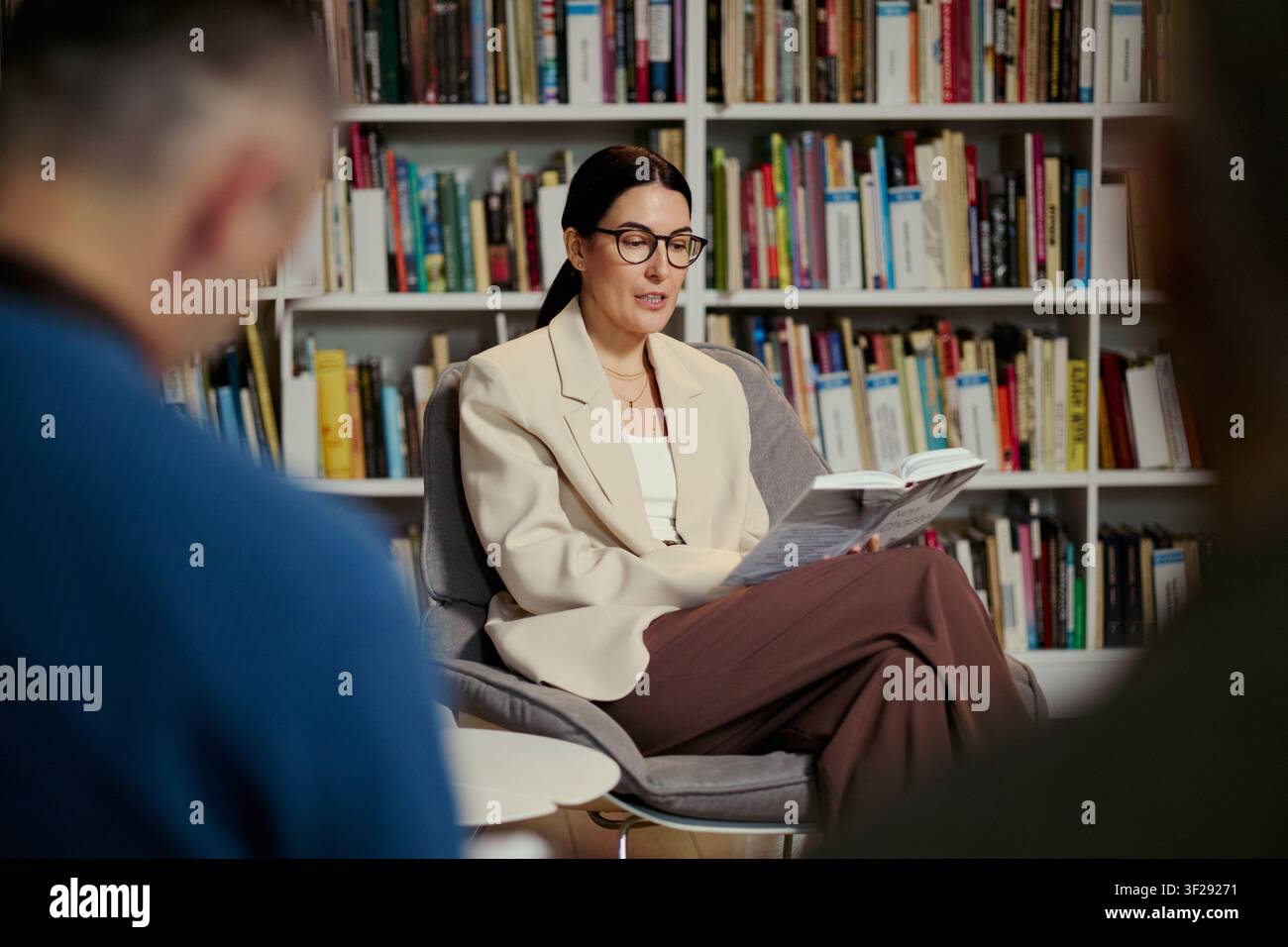 Donna caucasica di mezza età che legge da un libro durante una sessione di gruppo in biblioteca, guidando la discussione e l'apprendimento. Utile per educazione, coaching, alfabetizzazione, promozione di workshop Foto Stock