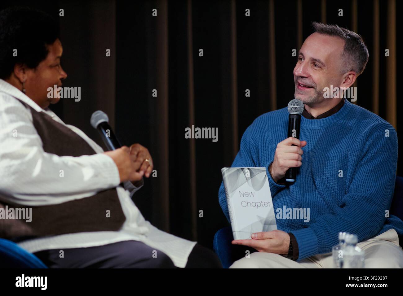 Scatto medio di relatori di mezza età che discutono di un libro durante un'intervista dal vivo, tenendo microfoni sul palco. Adatto per pubblicazioni, eventi per autori, campagne di comunicazione mediatica Foto Stock