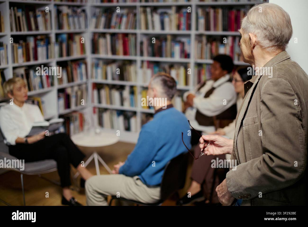 Uomo anziano in piedi e partecipa a una discussione di libro con un gruppo adulto diversificato in biblioteca, condividendo la sua prospettiva e imparando attivamente. Utile per l'educazione, la comunità, le campagne di alfabetizzazione Foto Stock