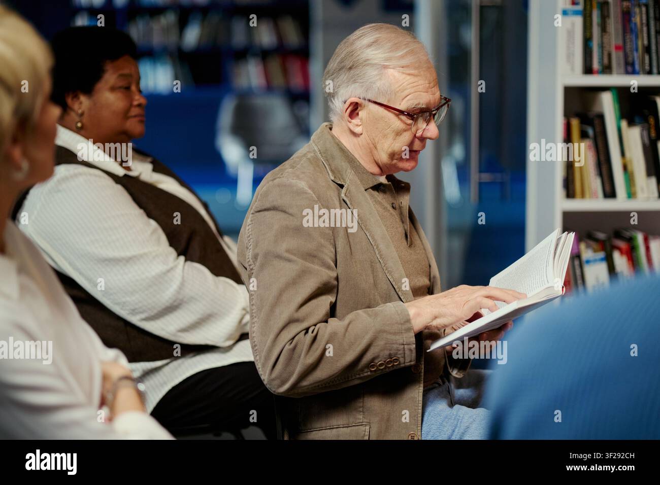 Uomo anziano caucasico che legge un libro durante una discussione di gruppo in biblioteca, condividendo conoscenza e apprendimento permanente. Adatto per educazione, programmi comunitari, campagne di alfabetizzazione per anziani Foto Stock