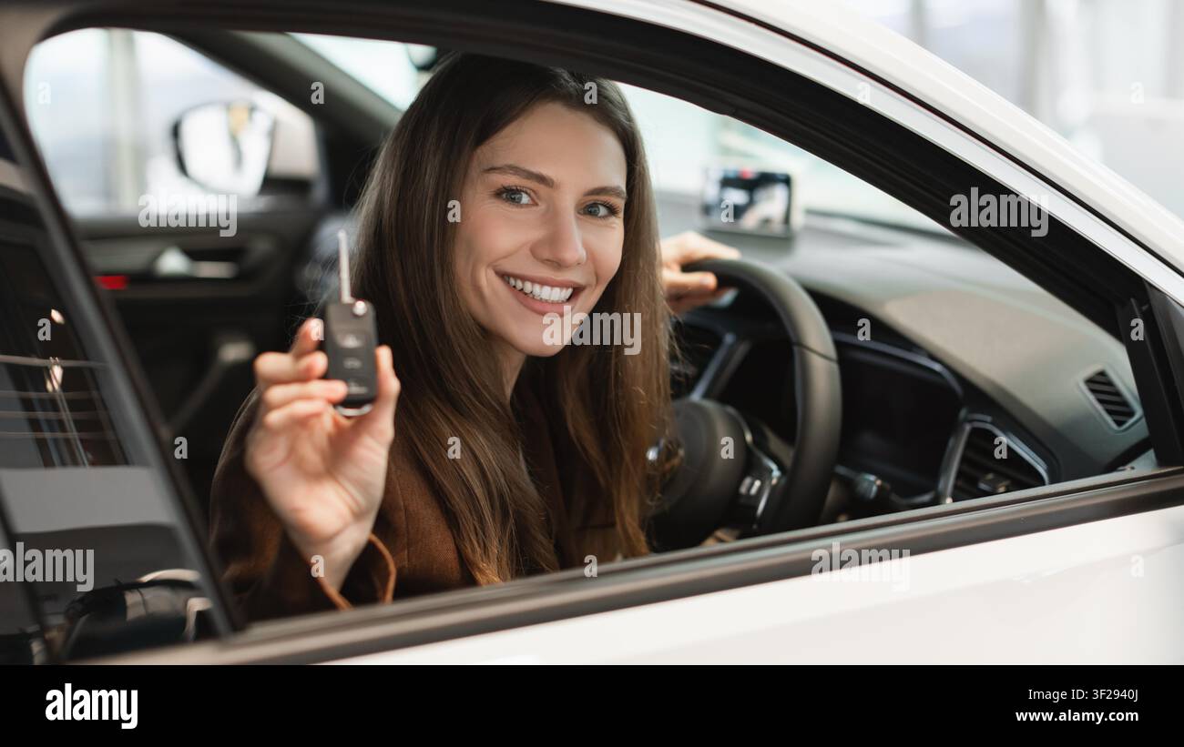 Giovane donna felice che guida un'auto nuova e tiene la chiave all'interno del veicolo, concetto di proprietà dell'auto Foto Stock